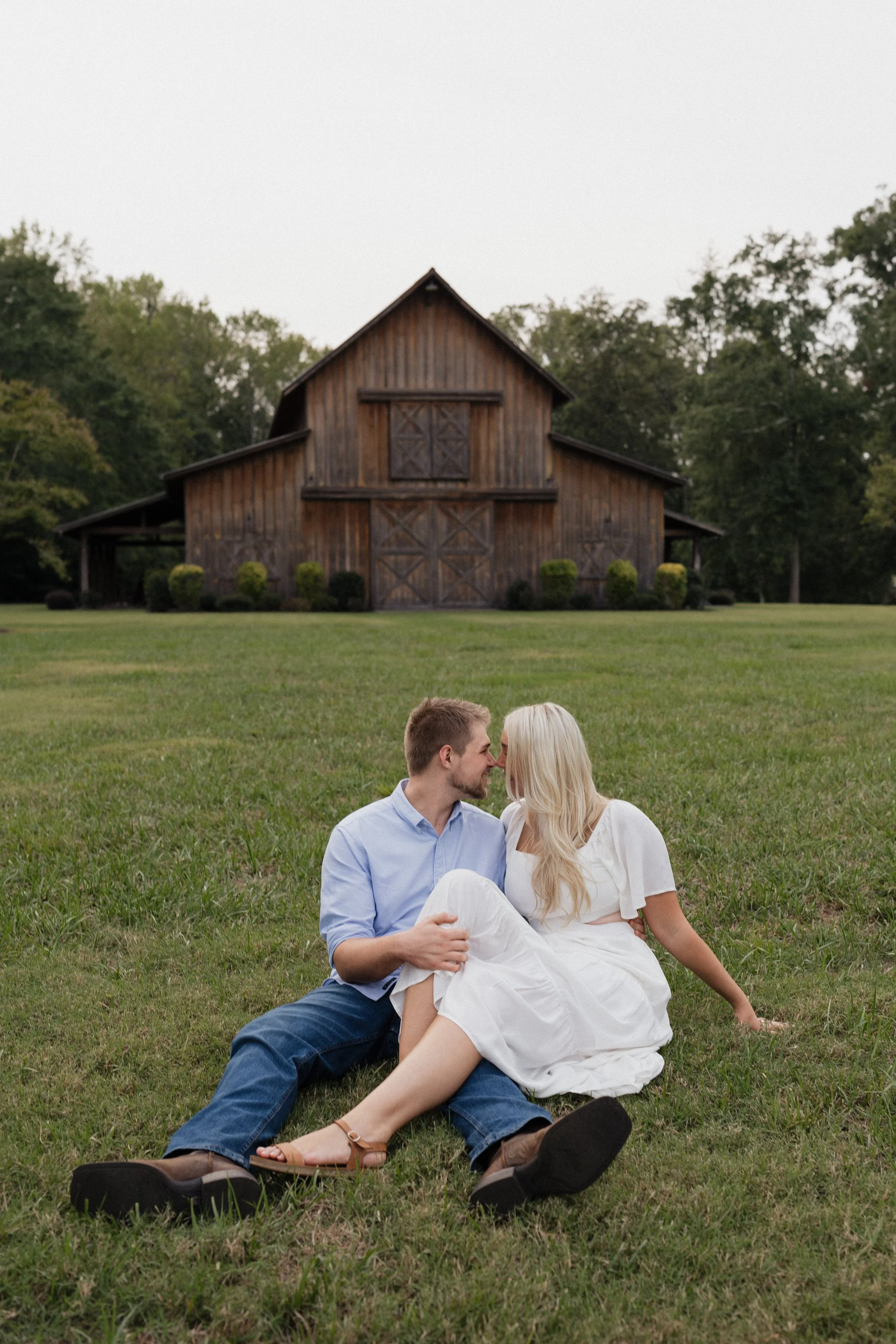 A couple sitting on the grass, about to kiss, with a rustic barn in the background.