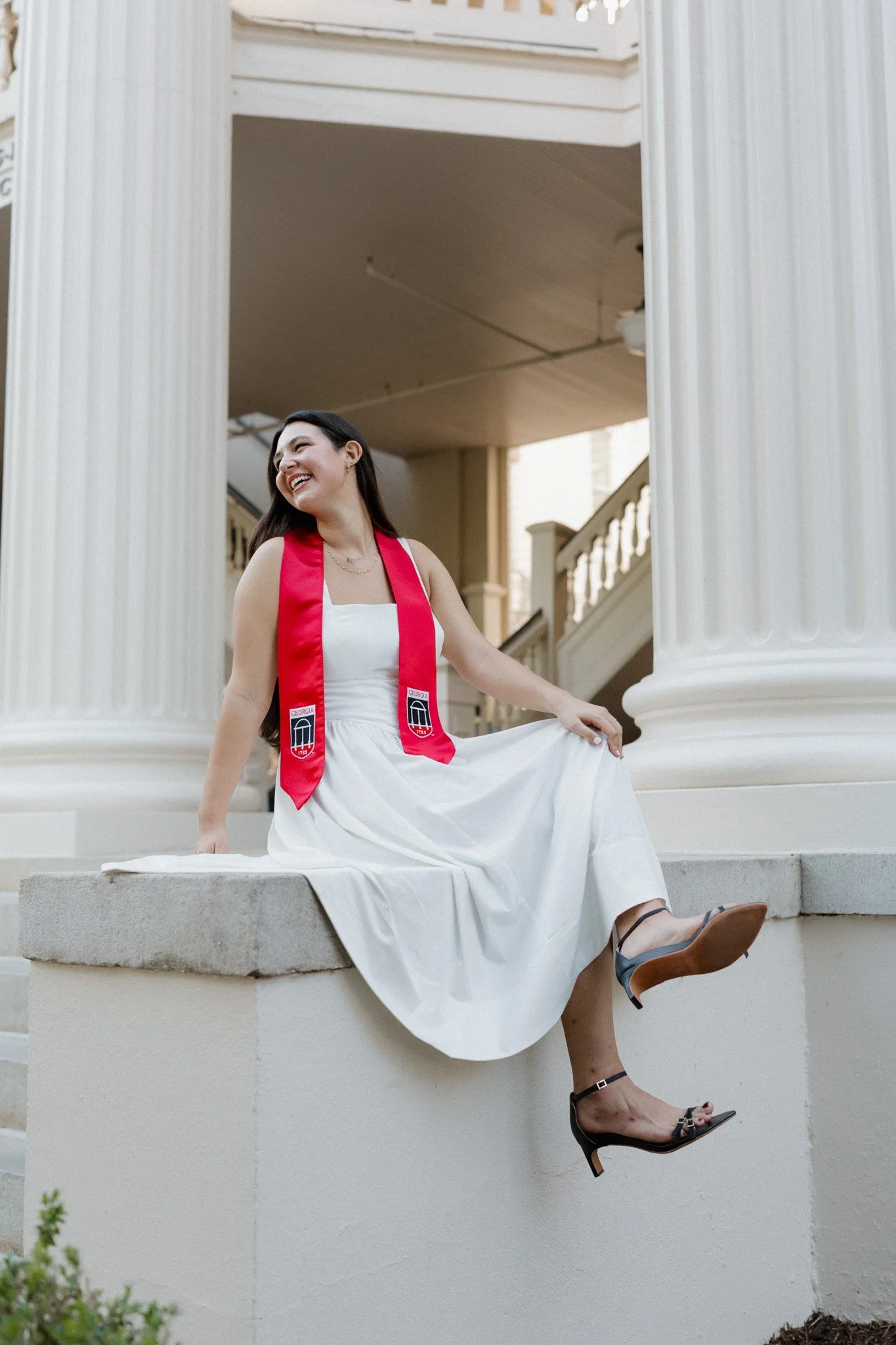 Young woman sitting on a ledge in front of large white columns, wearing a white dress, black heels, and a red graduation stole, smiling and looking to the side.