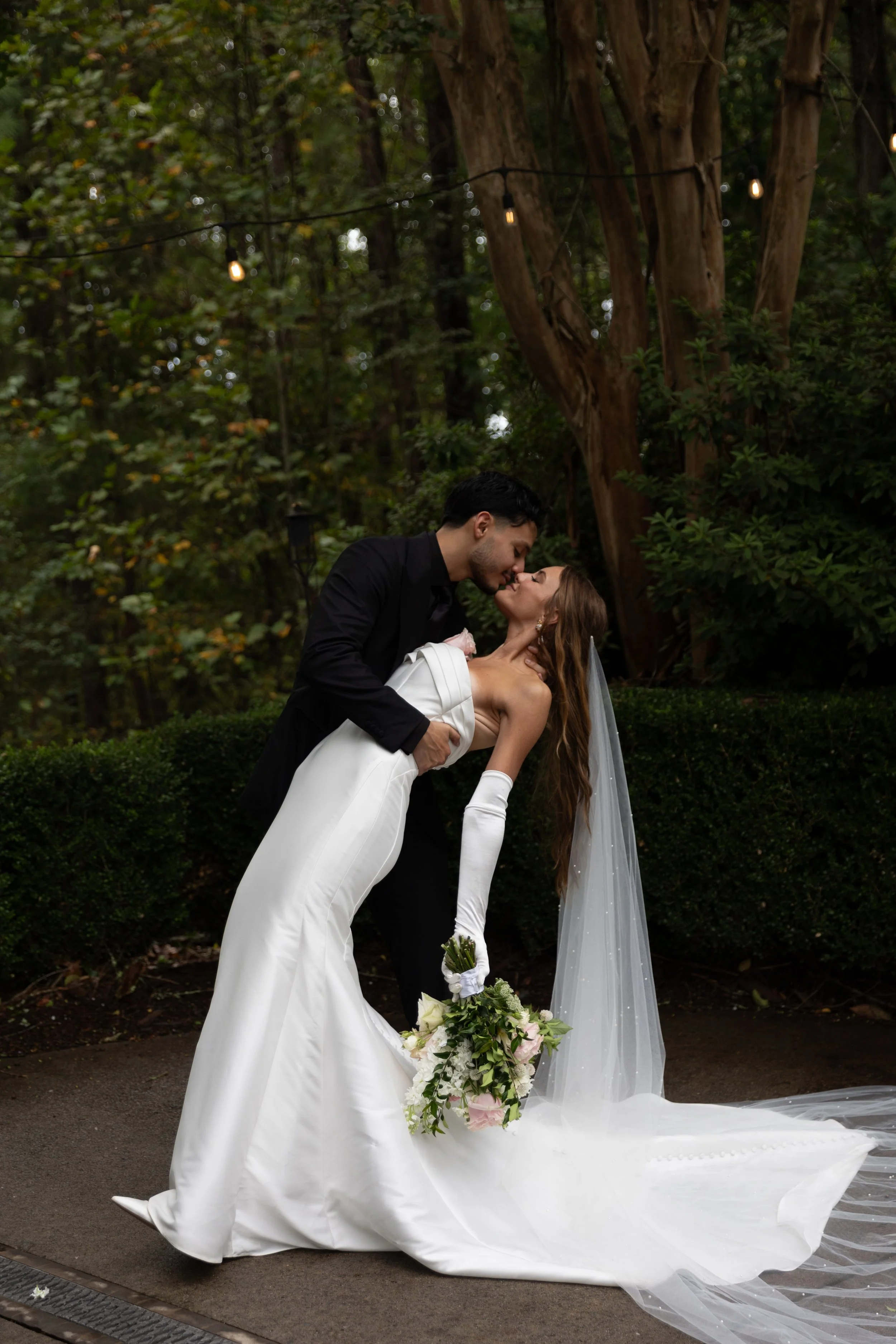 A bride in a white gown and long gloves and a groom in a black suit sharing a romantic kiss outdoors during their wedding, with trees and string lights in the background.