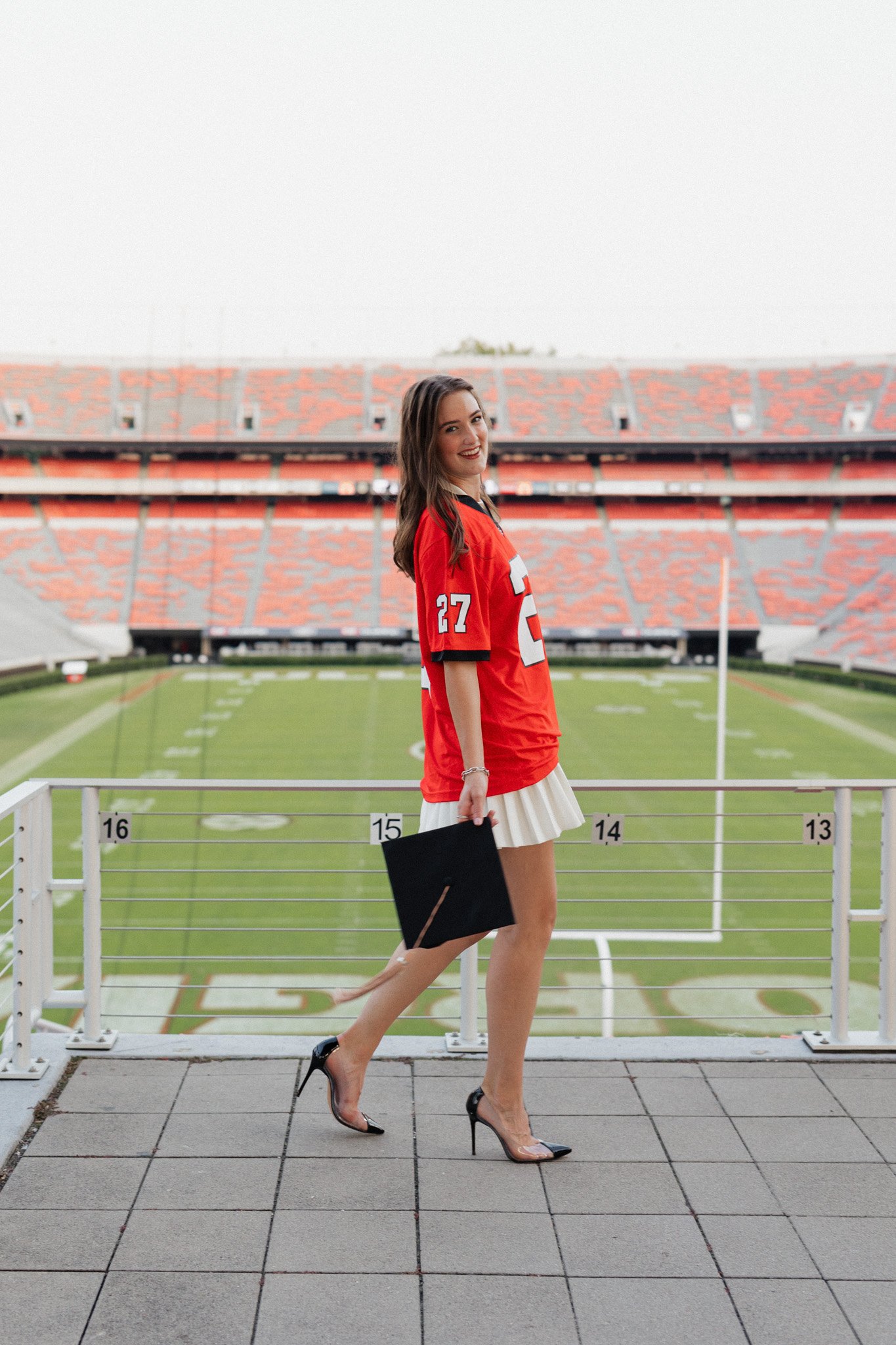A young woman in a red football jersey with the number 27, wearing a white pleated skirt and high heels, walking on a rooftop overlooking a football field, holding a black diploma cover and smiling at the camera.