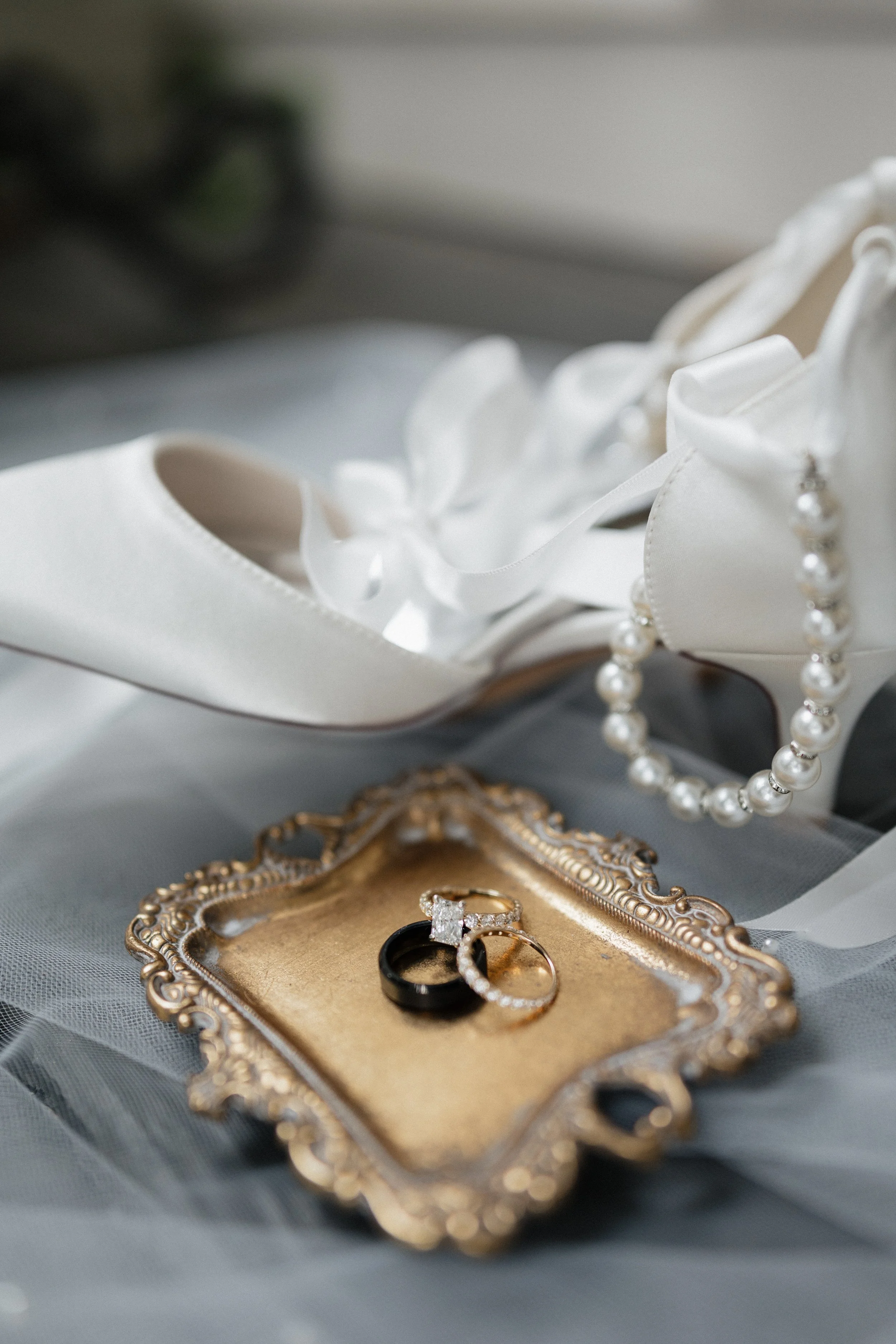 A pair of wedding rings, an engagement ring with a diamond, a black band, and a gold band, are placed on an ornate gold tray. A white satin wedding shoe with ribbon and flower details, and a pearl necklace, are in the background.