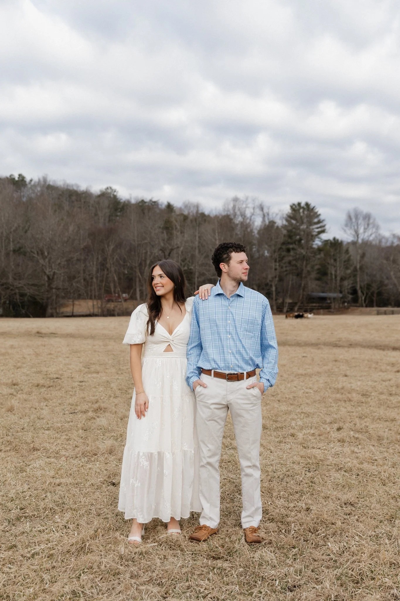 A young couple standing outdoors in a field on a cloudy day, the woman wearing a white dress and the man wearing a blue checked shirt and beige pants.