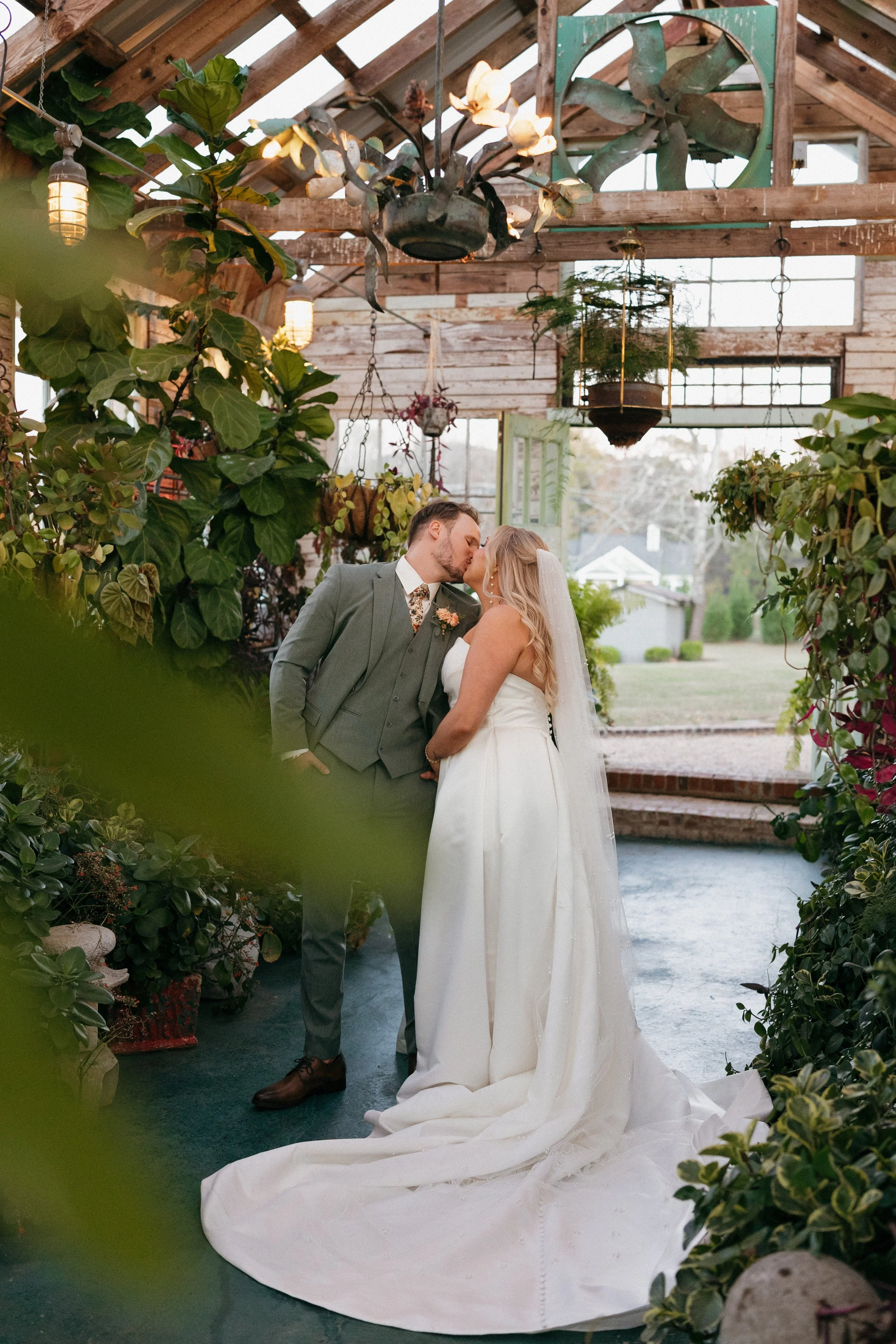 A bride and groom share a kiss in a lush, greenhouse-style setting with abundant green plants, hanging pots, and natural light during their wedding ceremony.