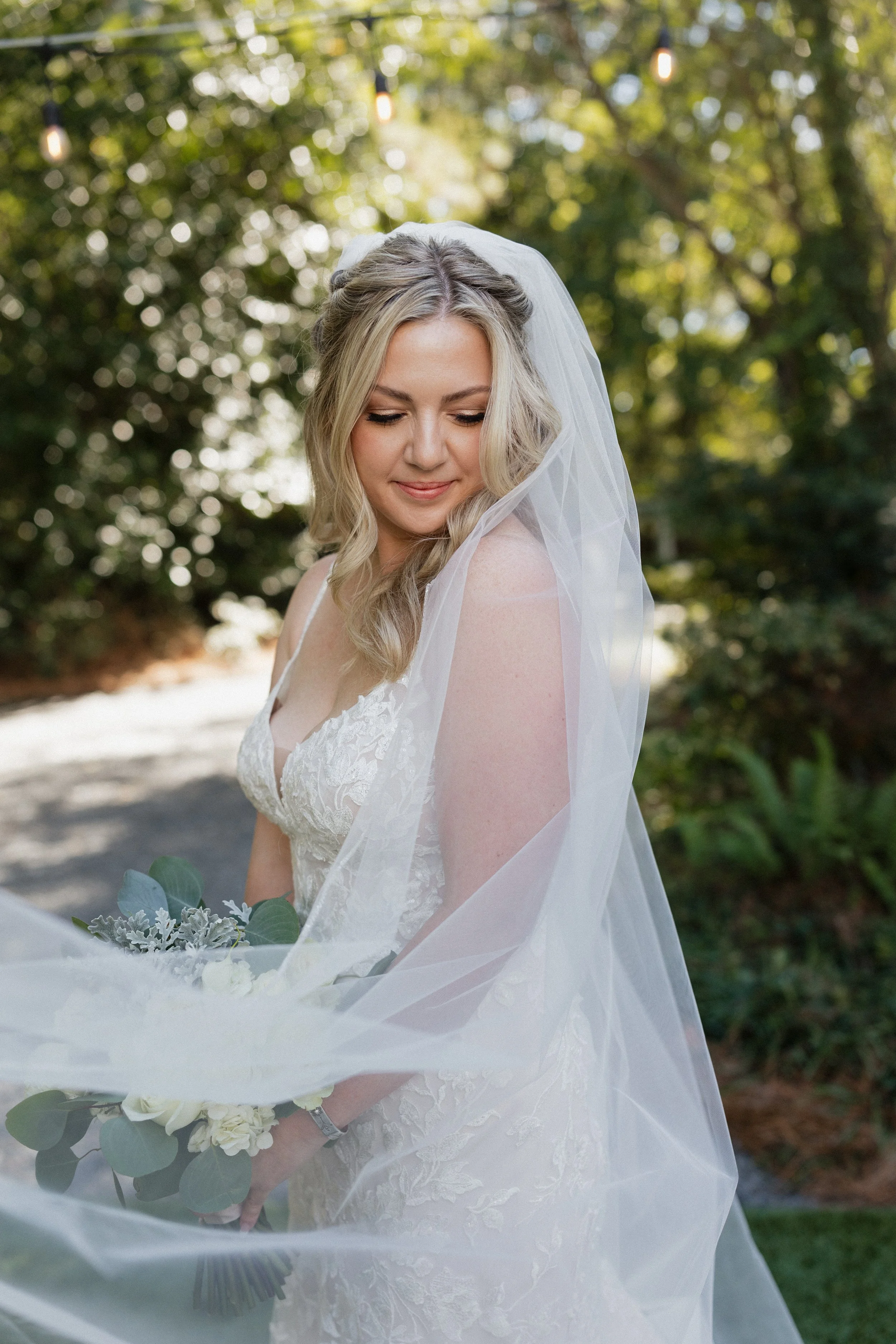 A bride in a white lace wedding dress and veil holding a bouquet of greenery and white flowers, standing outdoors with trees and sunlight in the background.