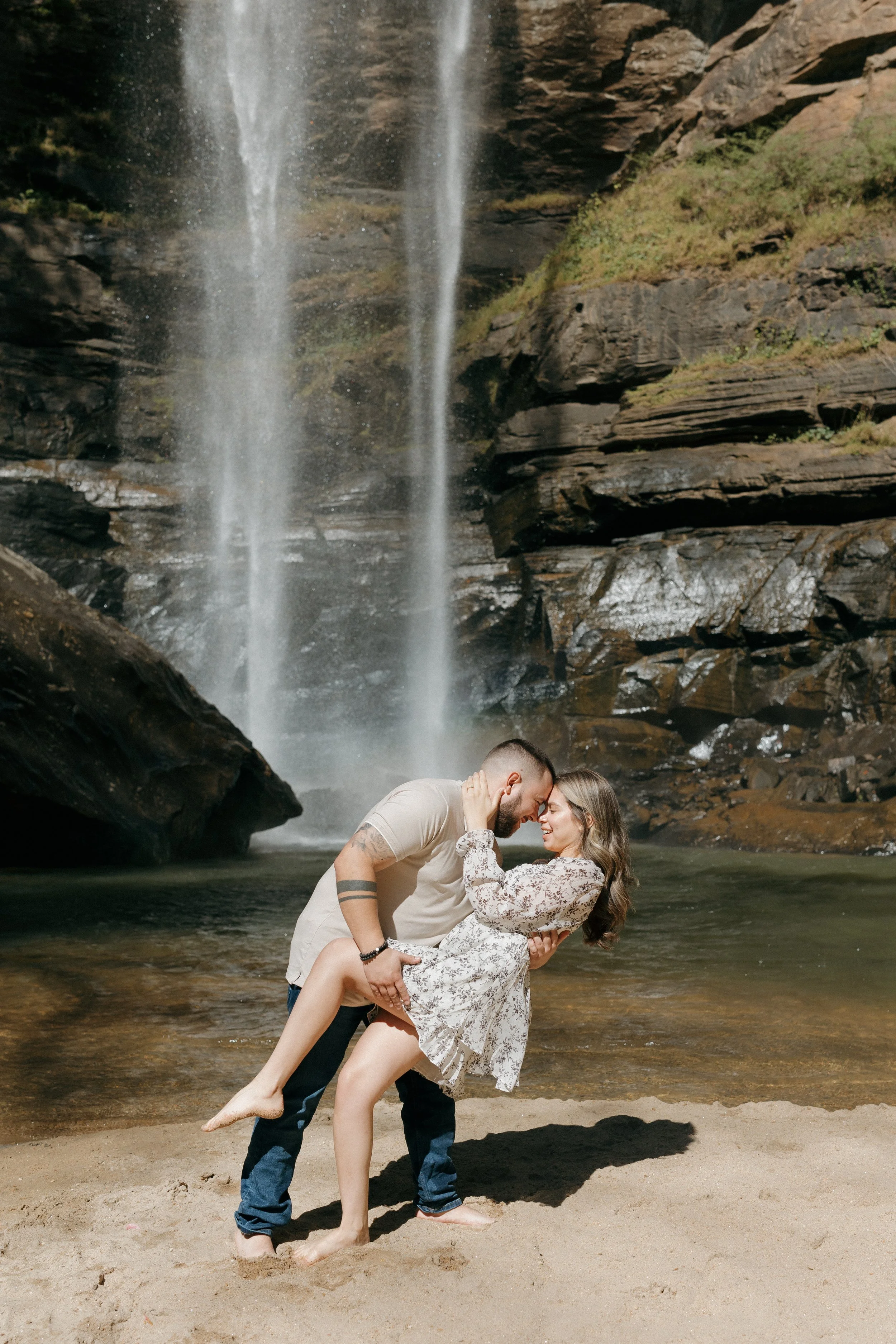 A couple dancing on the sandy beach in front of a waterfall, with water cascading down rocks in the background.