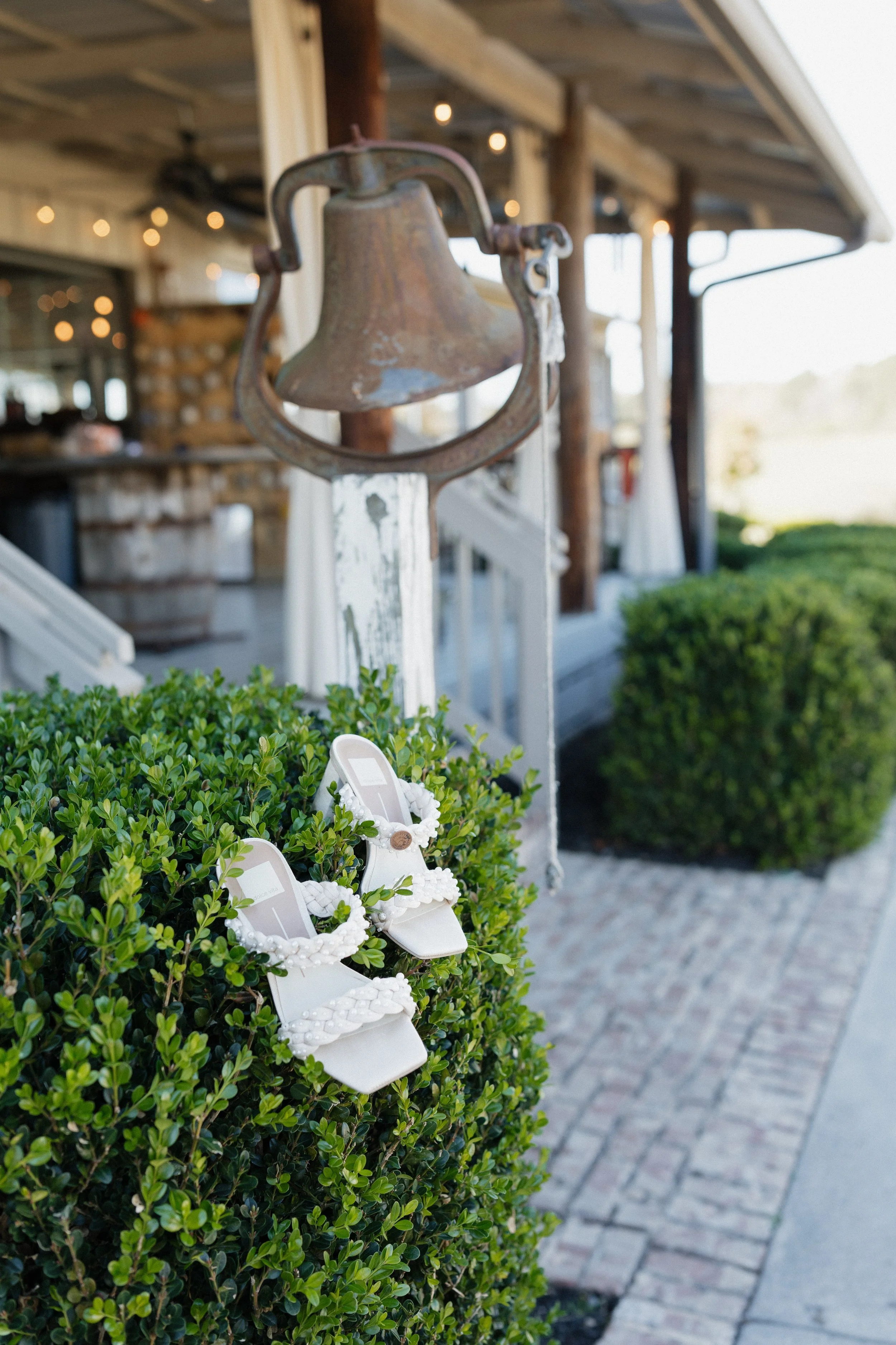 A pair of white sandals with decorative straps resting on a green shrub outside a rustic building with string lights and wooden elements.