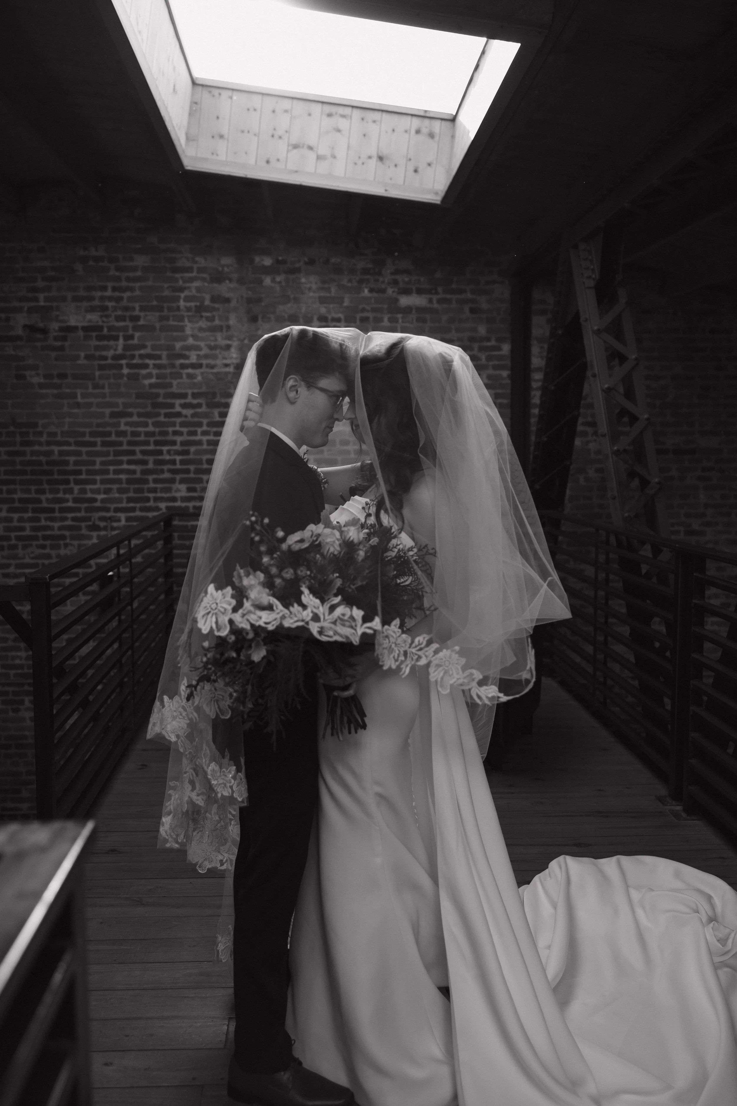 A bride and groom in a black and white photo, standing close together under a veil, with the bride holding a bouquet of flowers, in an indoor setting with brick walls and a skylight overhead.