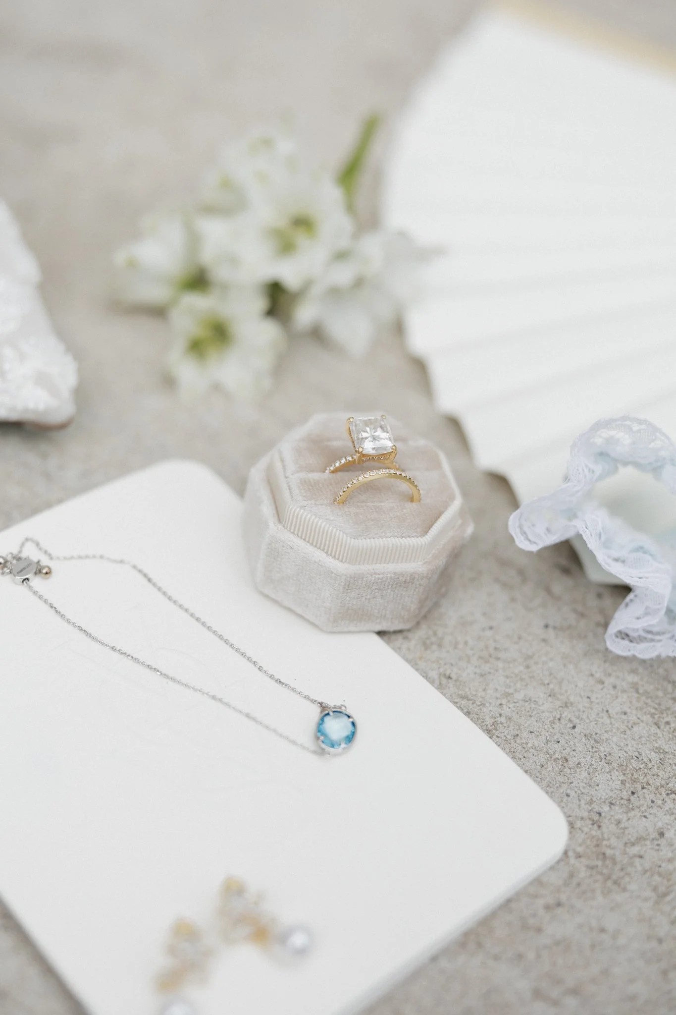 Close-up of jewelry including a ring with a large clear gemstone in a velvet ring box, a delicate necklace with a blue gemstone, and a pair of earrings, all displayed on a white surface with white flowers and lace accessories in the background.