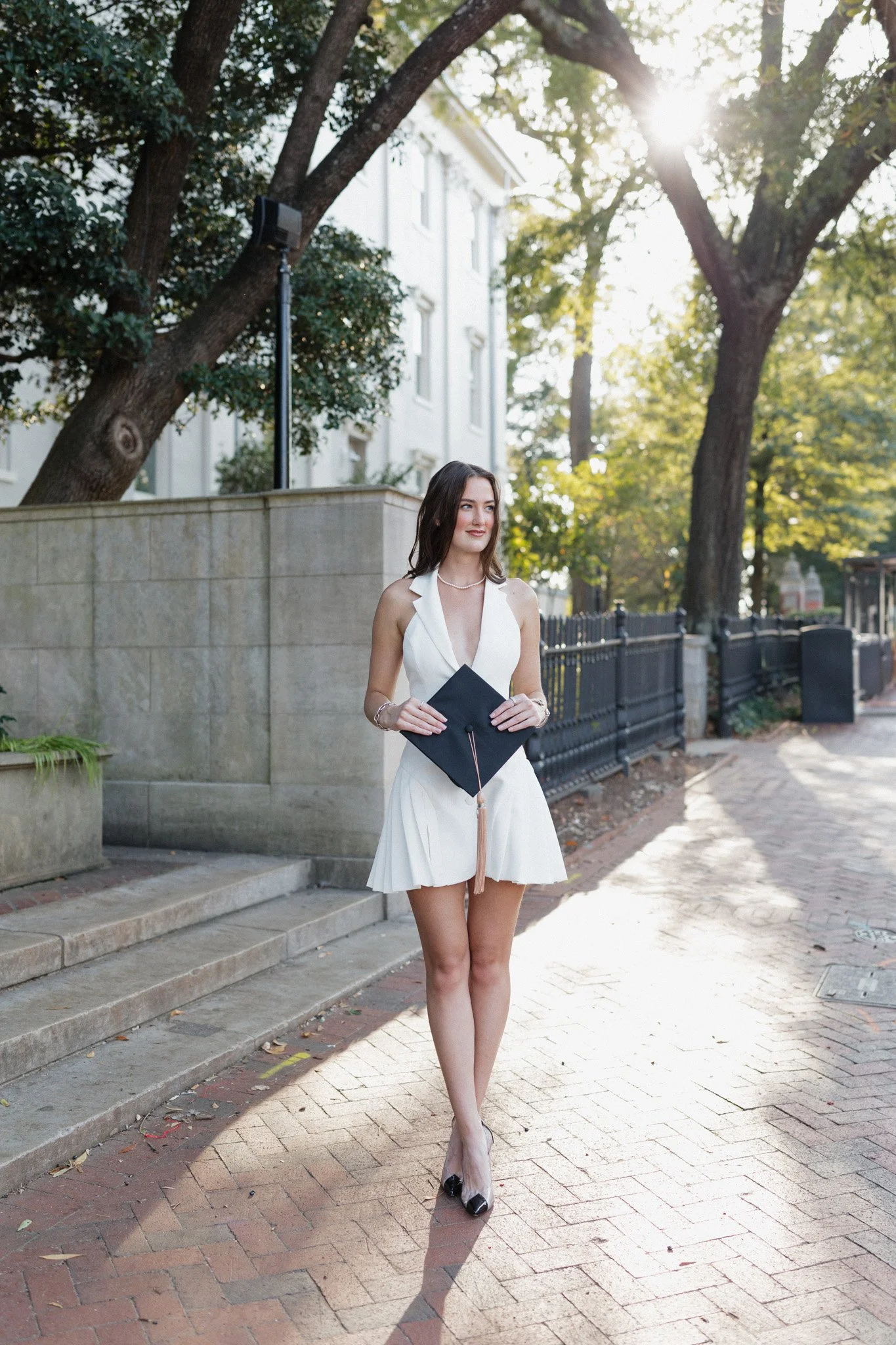 A young woman in a white dress holding a black graduation cap standing on a brick sidewalk outdoors in the evening with trees and a building in the background.