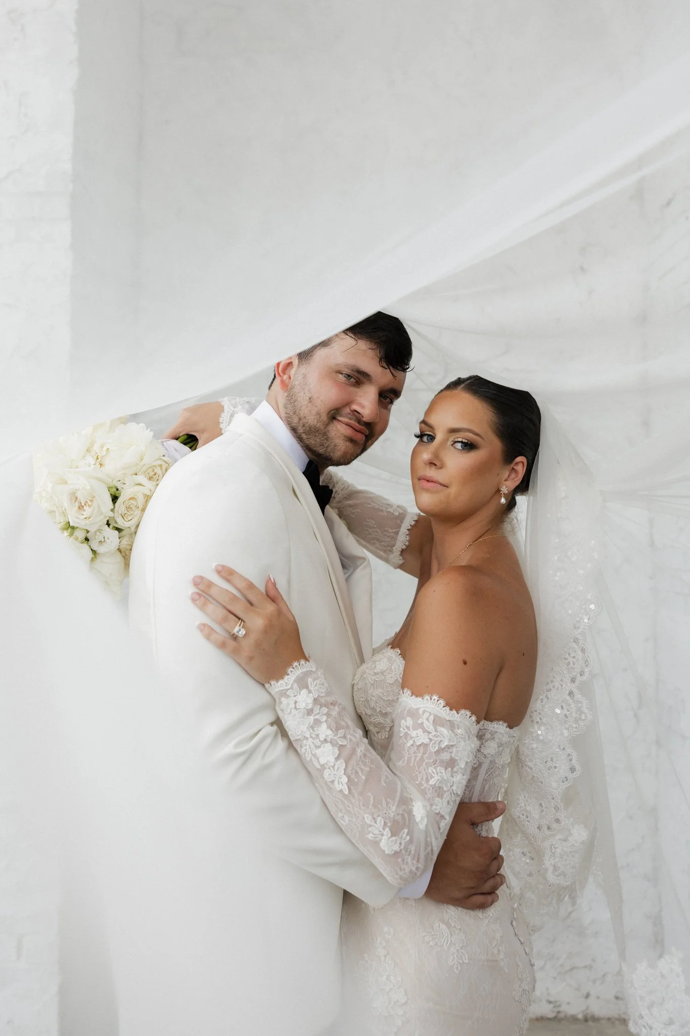 A bride and groom holding each other under a white wedding veil, with the bride in a lace wedding dress and the groom in a white suit, standing in front of a white brick wall.