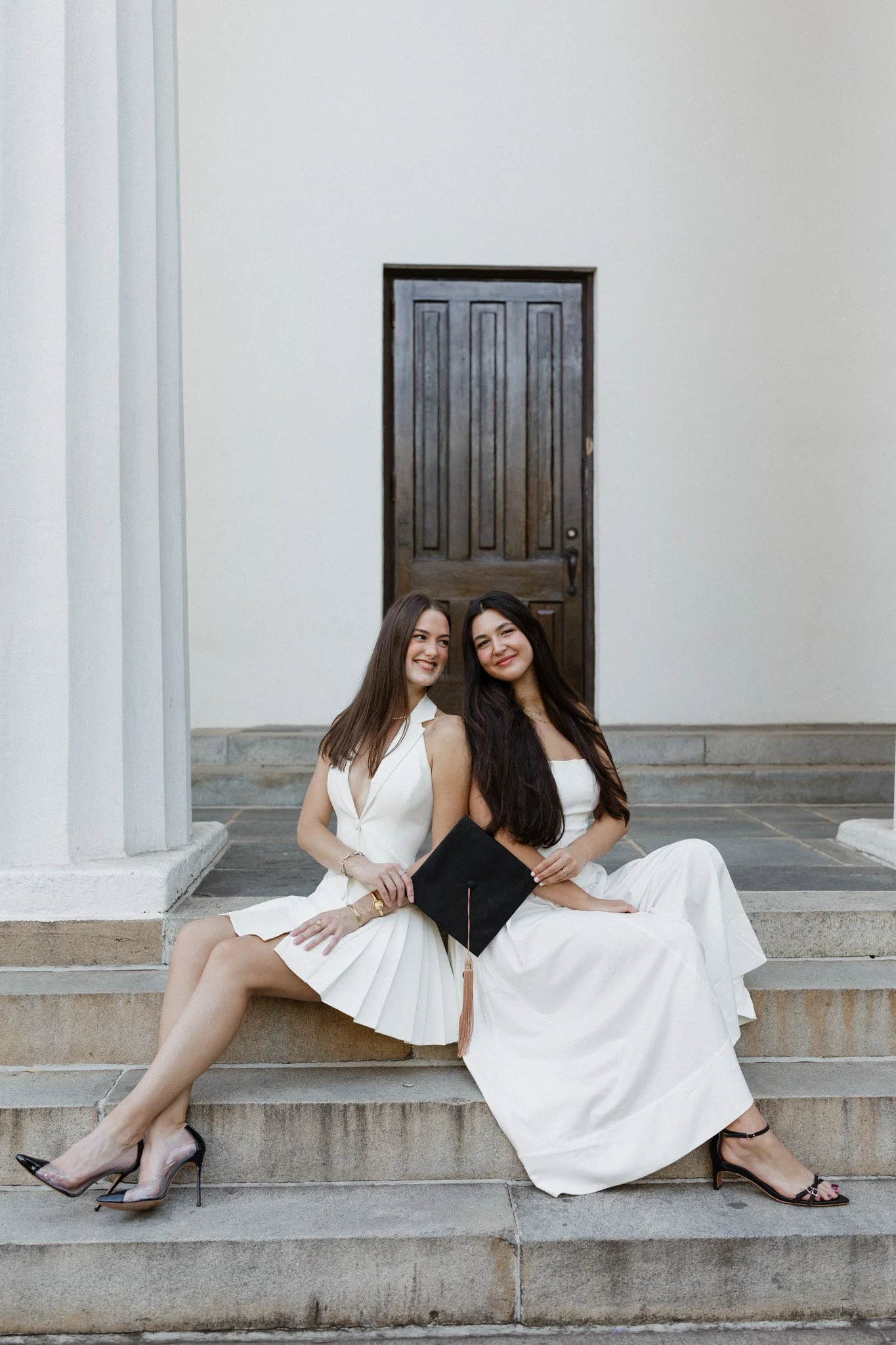 Two young women dressed in white sitting on steps in front of a dark wooden door, one holding a black graduation cap.