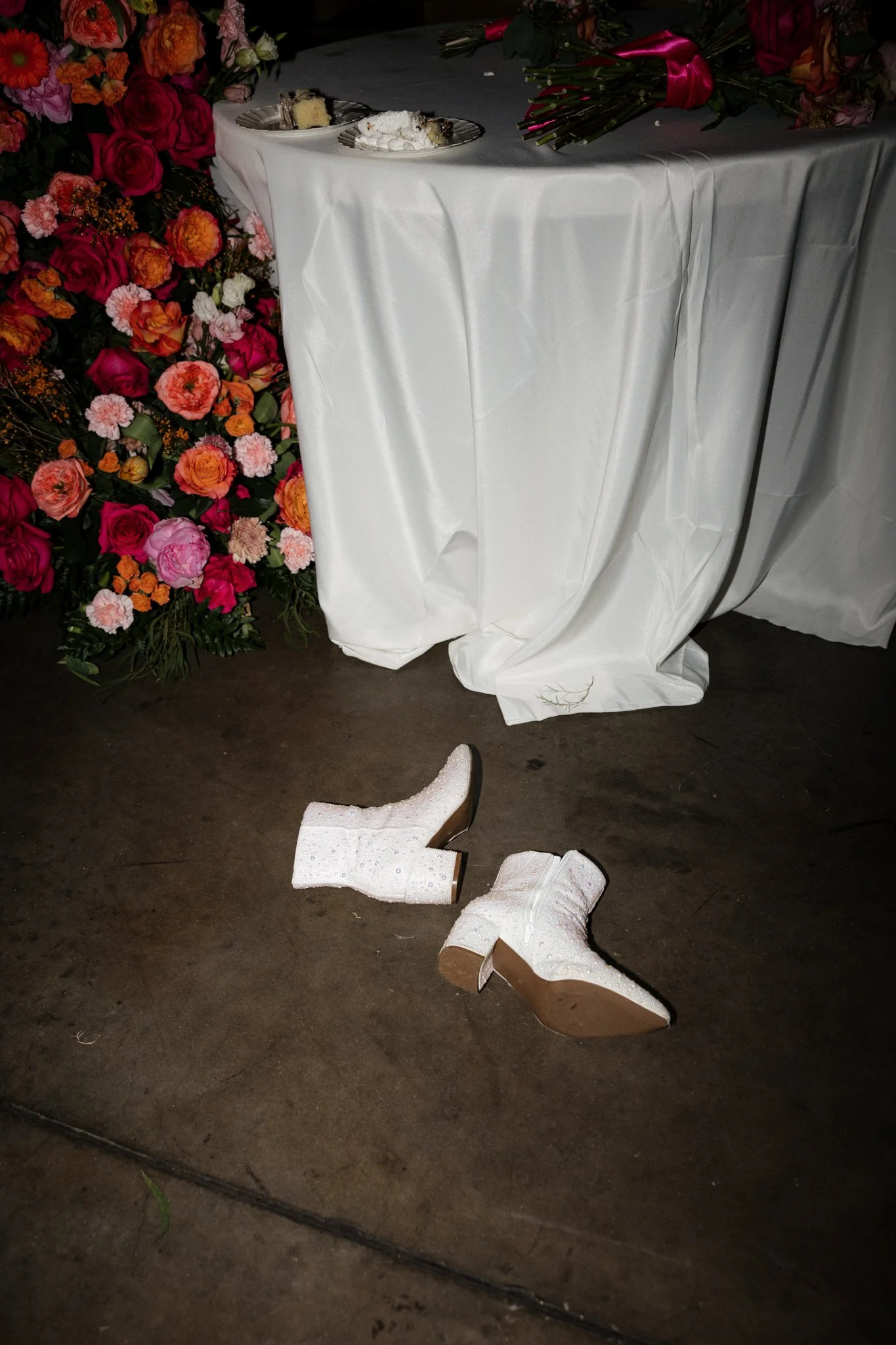 White cowboy boots on a dark floor near a decorated table with flowers and overturned plates; the boots are sparkly and appear to have fallen.