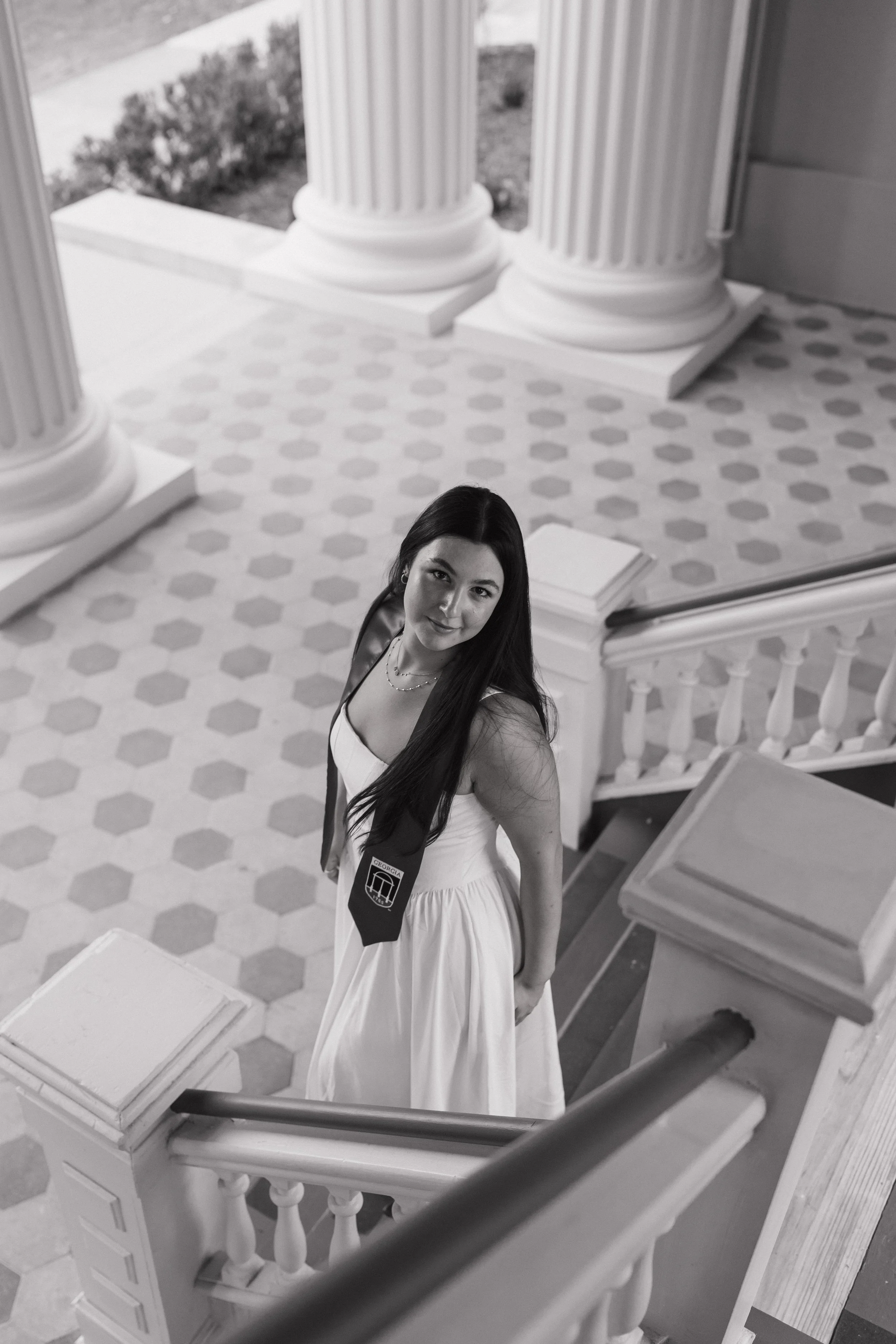 A young woman with long dark hair and a white dress standing on a staircase inside a building with large columns, looking up at the camera with a slight smile.