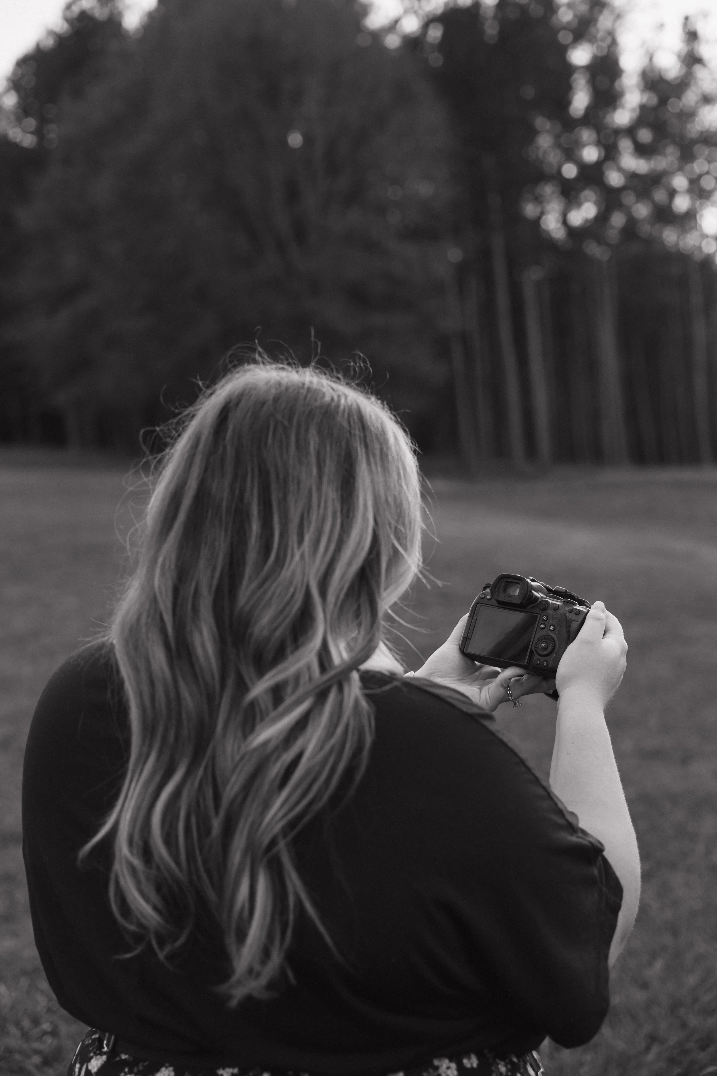 A woman with wavy hair taking a picture with a digital camera outdoors in a natural setting, with trees and grass in the background.