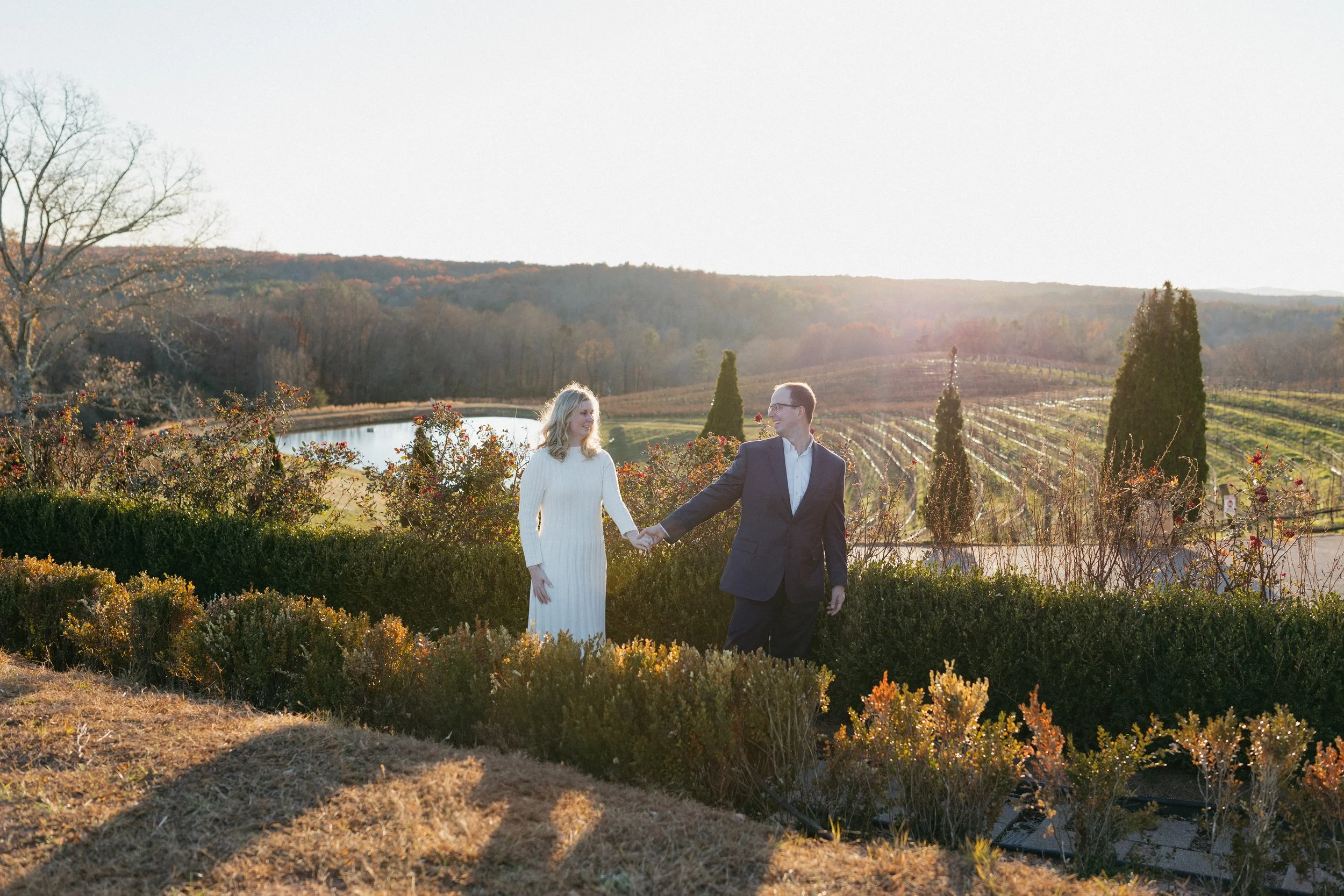 A couple holding hands and walking outdoors in a scenic vineyard during sunset.