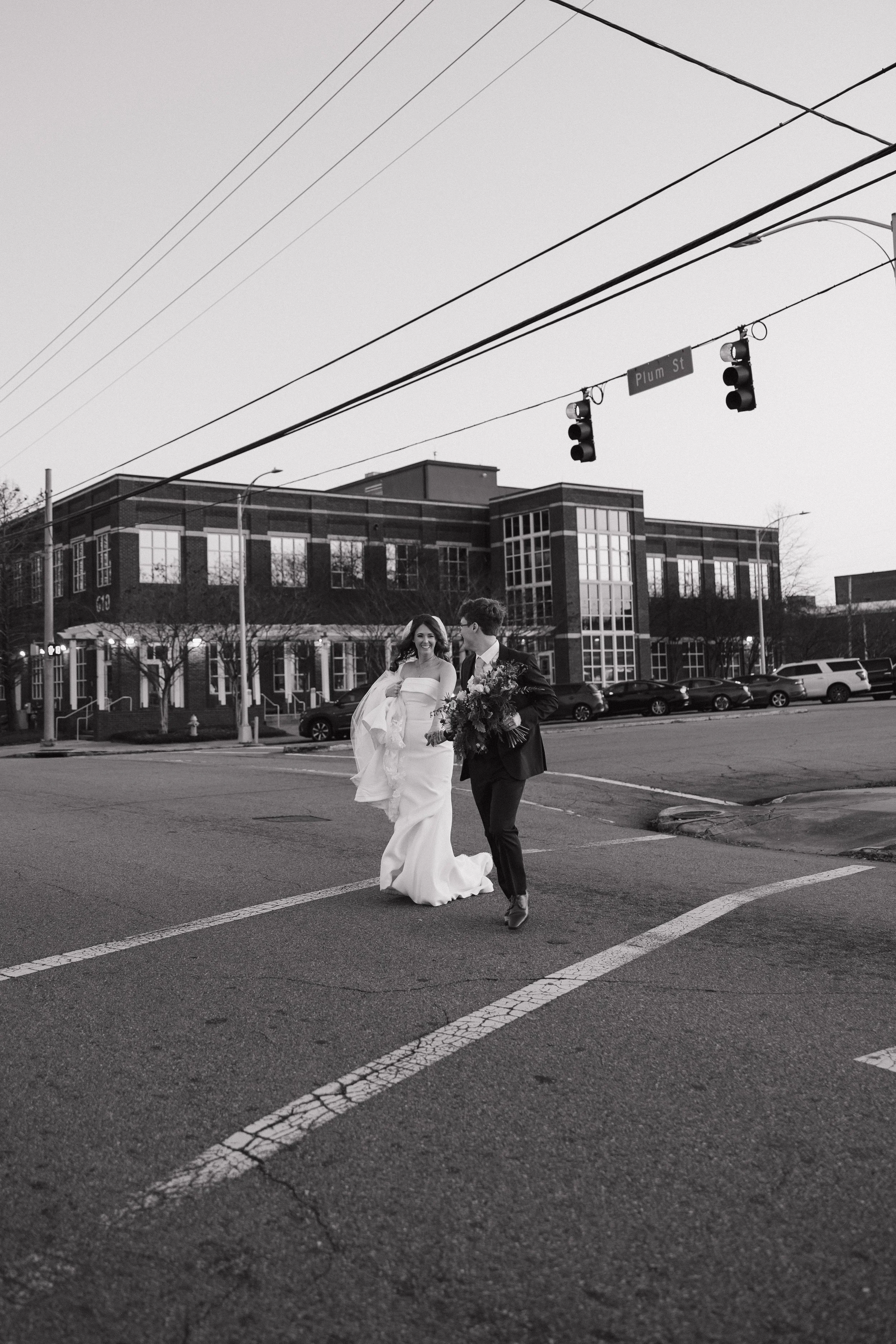 A black and white photo of a bride and groom walking across a city street, with the bride wearing a wedding dress and veil, and the groom in a dark suit holding a bouquet of flowers, under a traffic light and street sign that reads "Plum St." In the background, there is a modern brick building and parked cars.