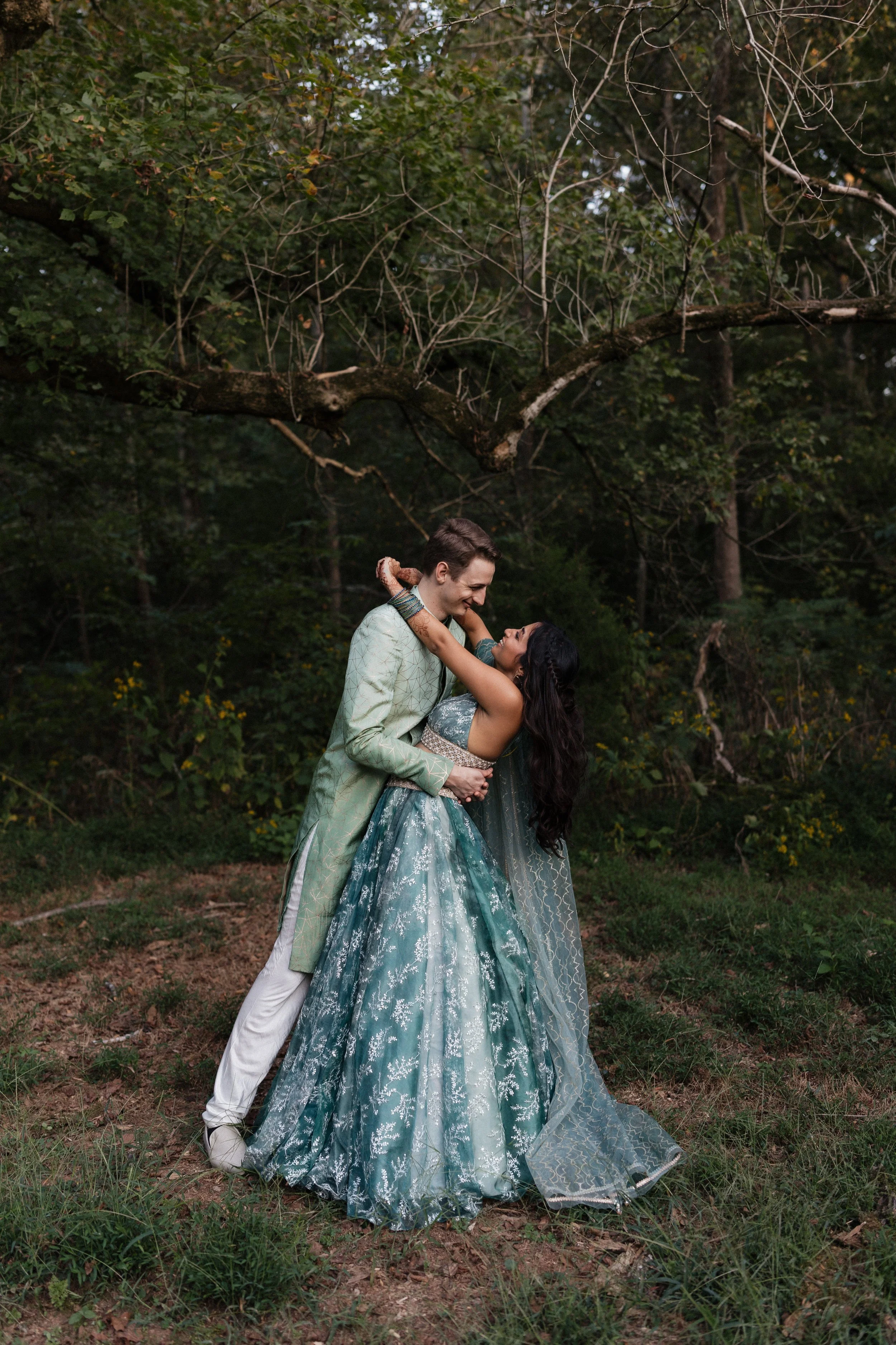 A couple wearing traditional Indian attire dancing outdoors in a forested area, holding each other and smiling.