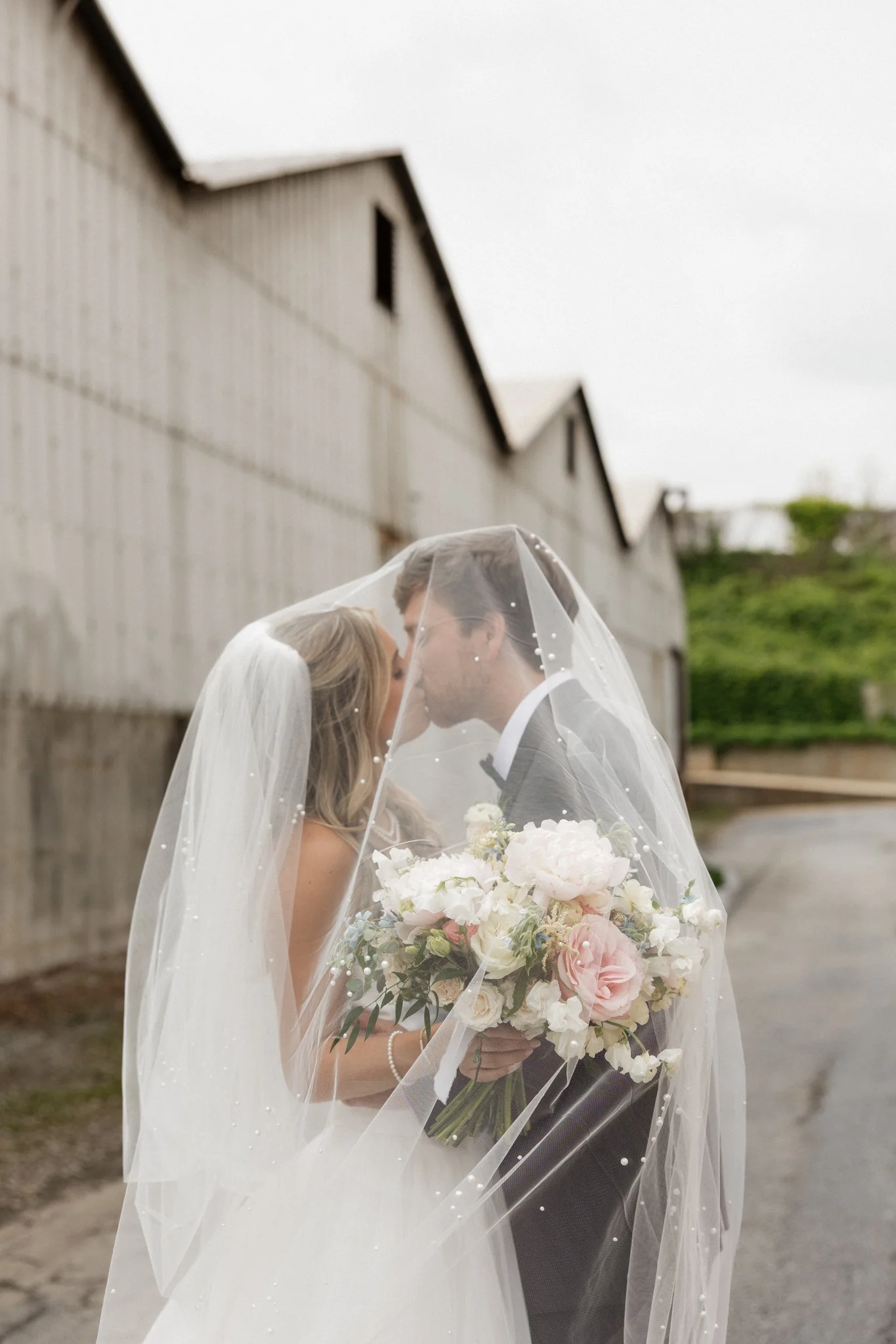 Bride and groom under a veil sharing a close moment, with the bride holding a large bouquet of pink and white flowers outside near warehouses.