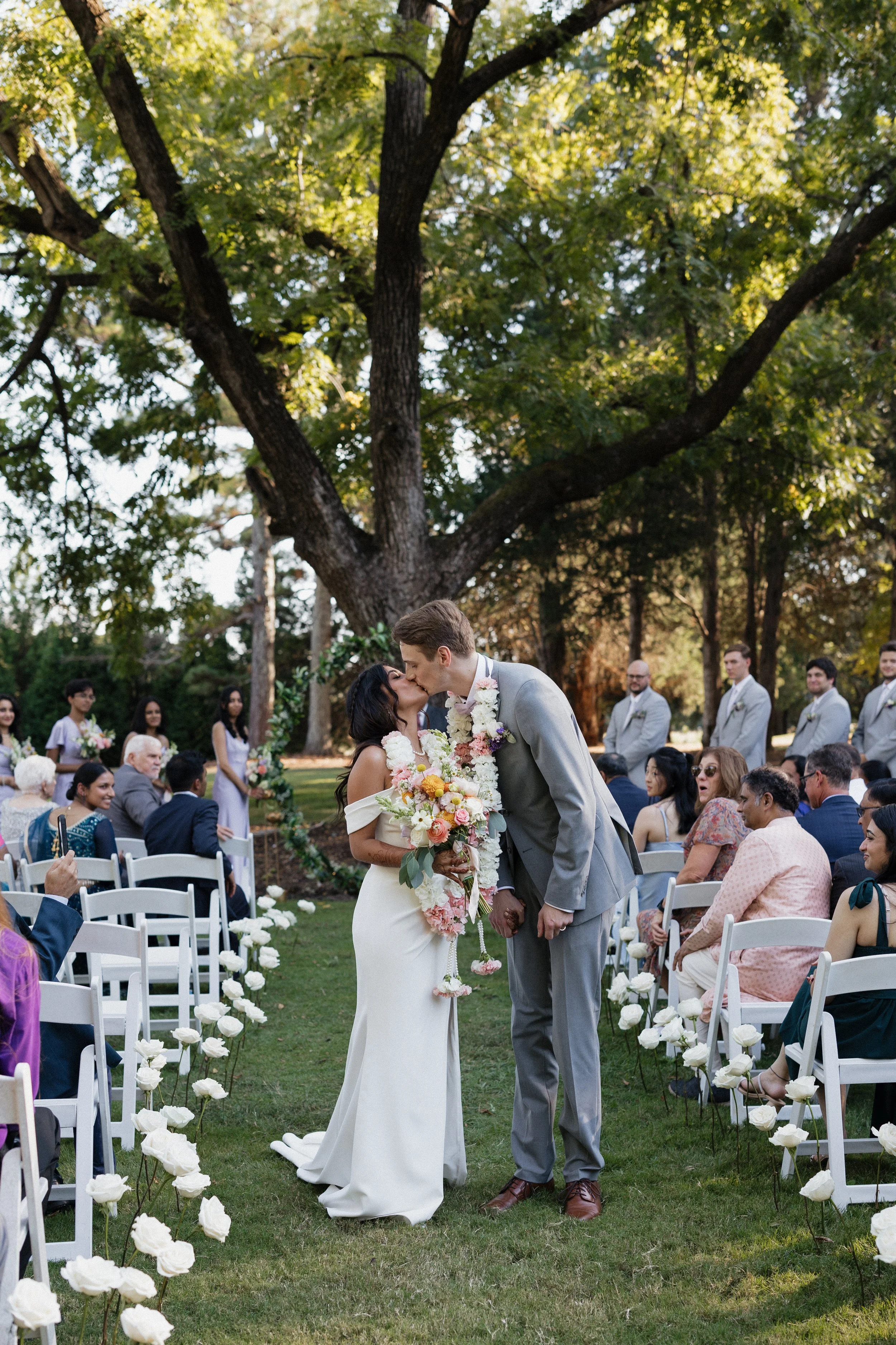 A bride and groom sharing a kiss during their outdoor wedding ceremony under a large tree, surrounded by seated guests and wedding party members on a grassy lawn decorated with white flowers.