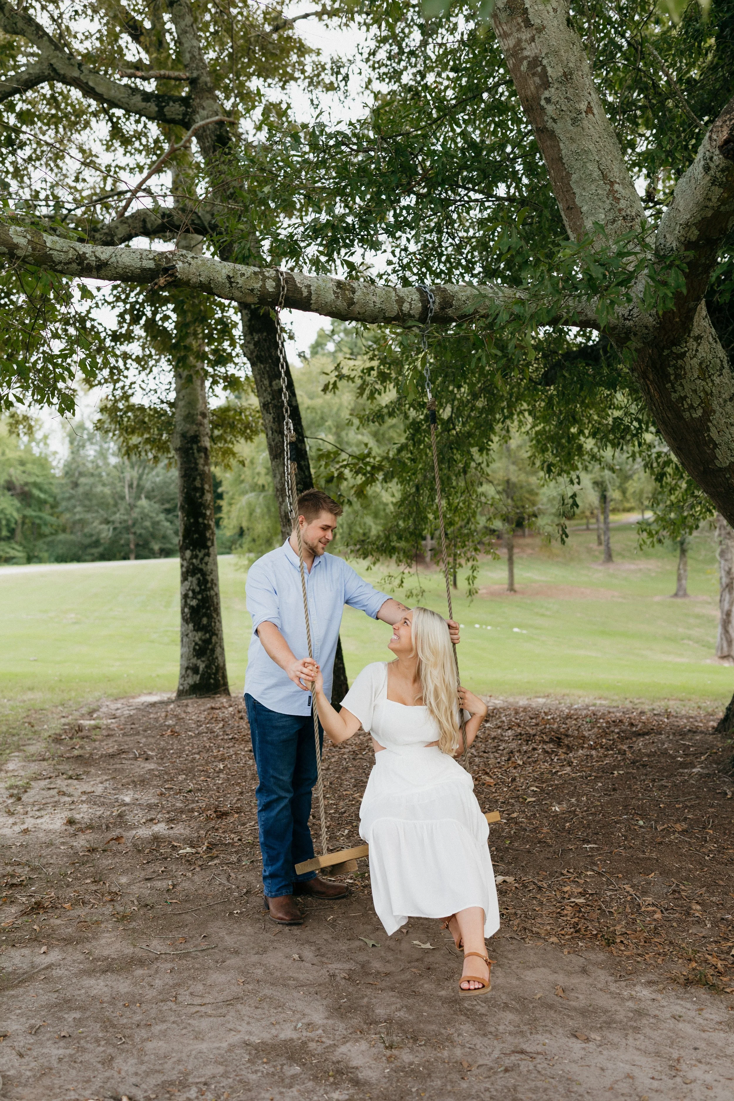 A man pushes a woman on a wooden swing hanging from a large tree in a park.