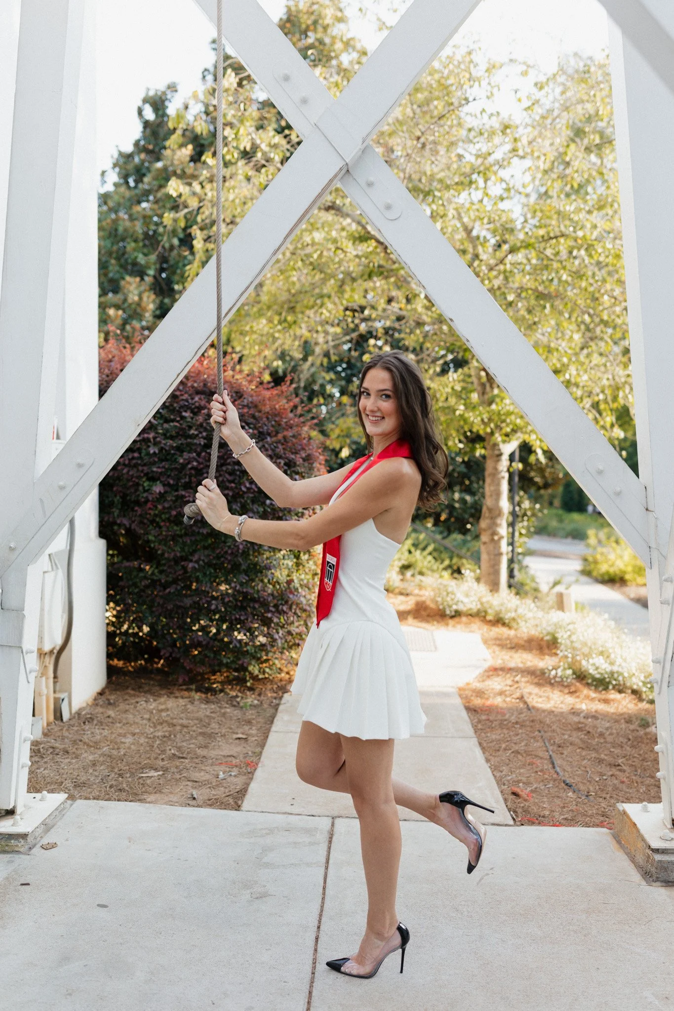 Young woman smiling in white dress, red tie, and high heels holding onto a swing under a white archway outdoors with trees and bushes in the background.