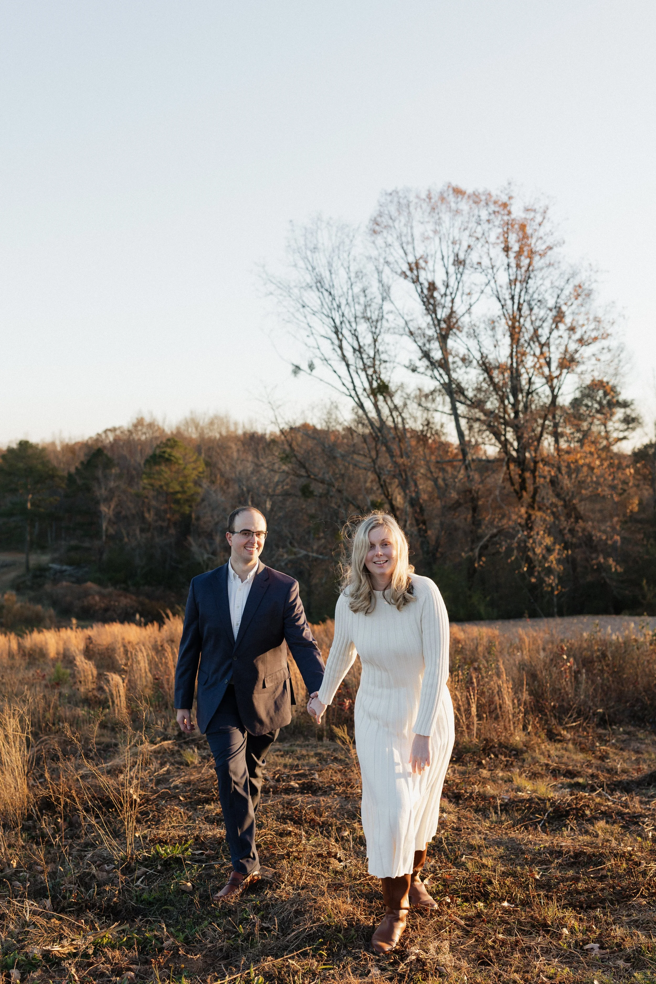 A man and woman walking hand-in-hand outdoors during sunset, surrounded by autumn trees and grass.