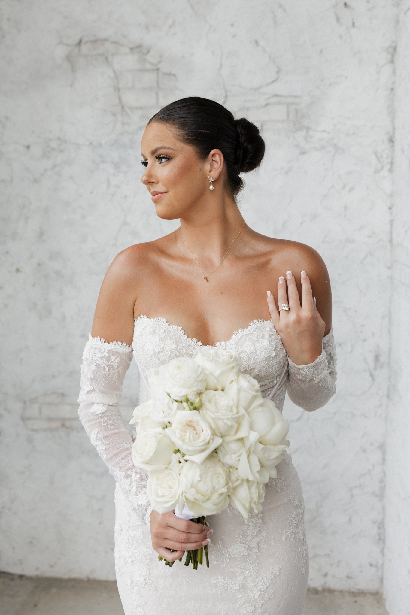 Bride holding a bouquet of white roses and calla lilies, wearing an off-the-shoulder lace wedding dress with long lace sleeves and diamond jewelry, standing against a white textured wall.