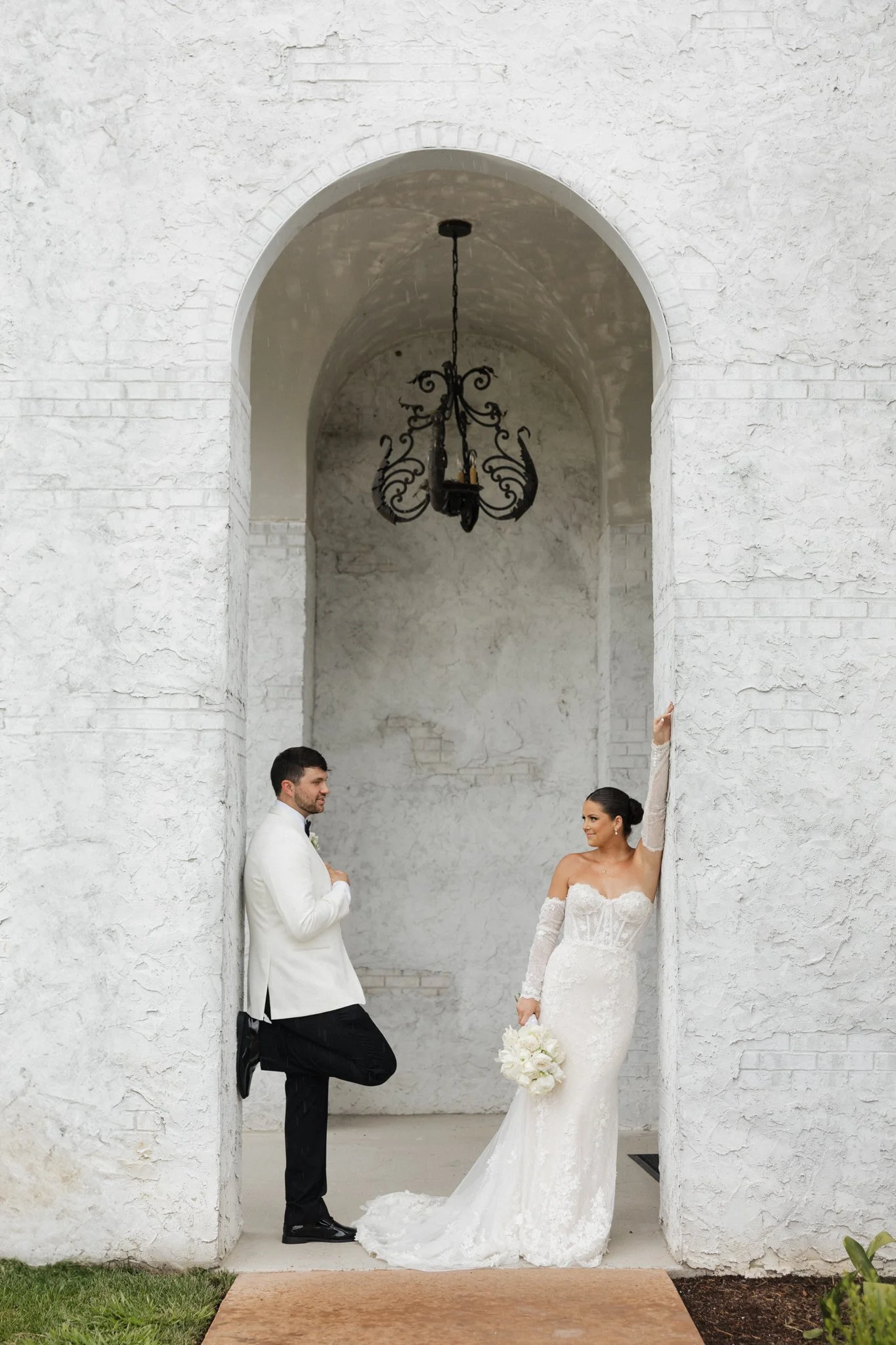 A bride and groom in wedding attire are posing in front of a white textured wall with an archway and black chandelier. The bride is holding a bouquet, and the groom is leaning against the wall.
