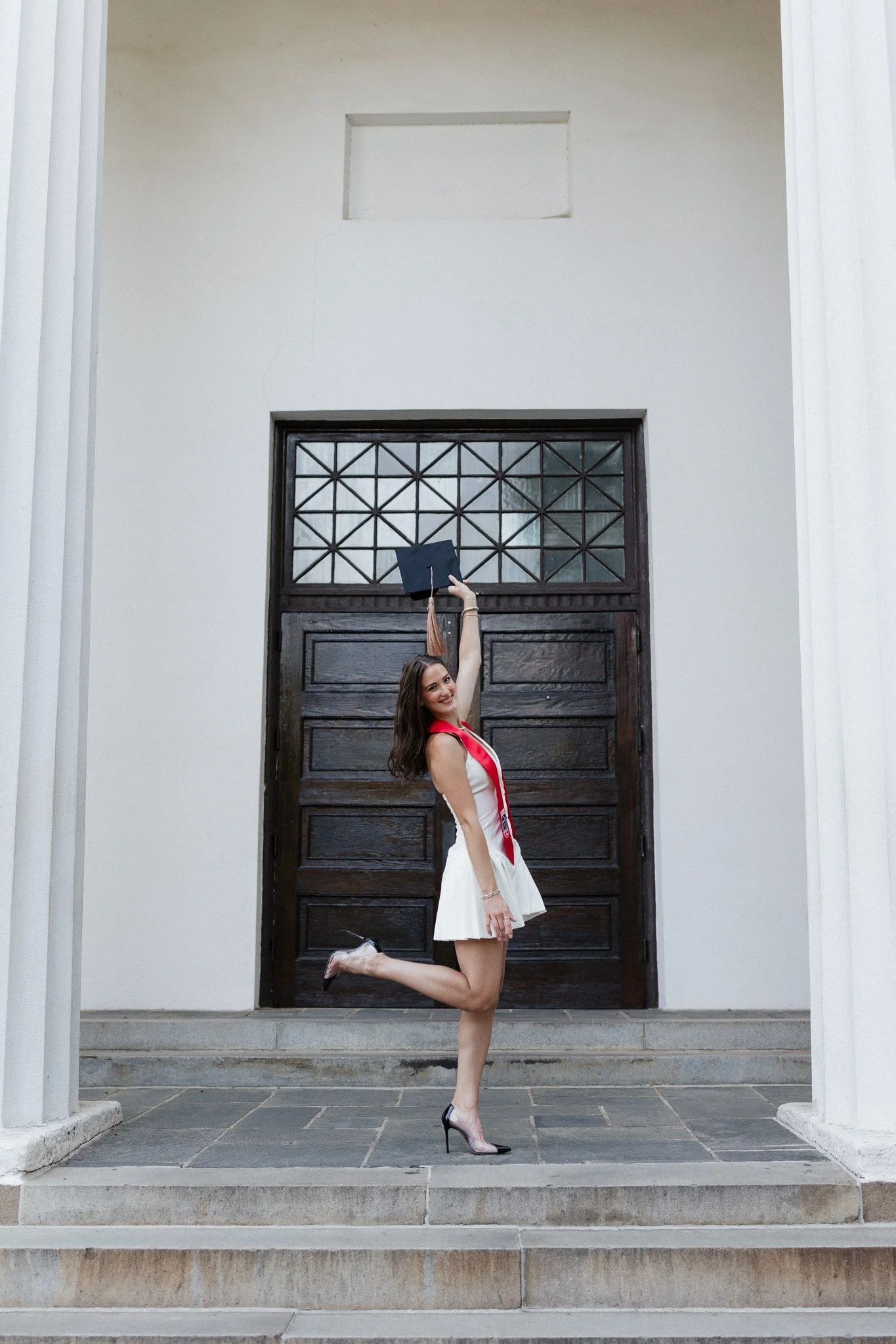 A young woman in a white dress and high heels celebrating graduation on the steps in front of a large wooden door, holding a graduation cap in the air.