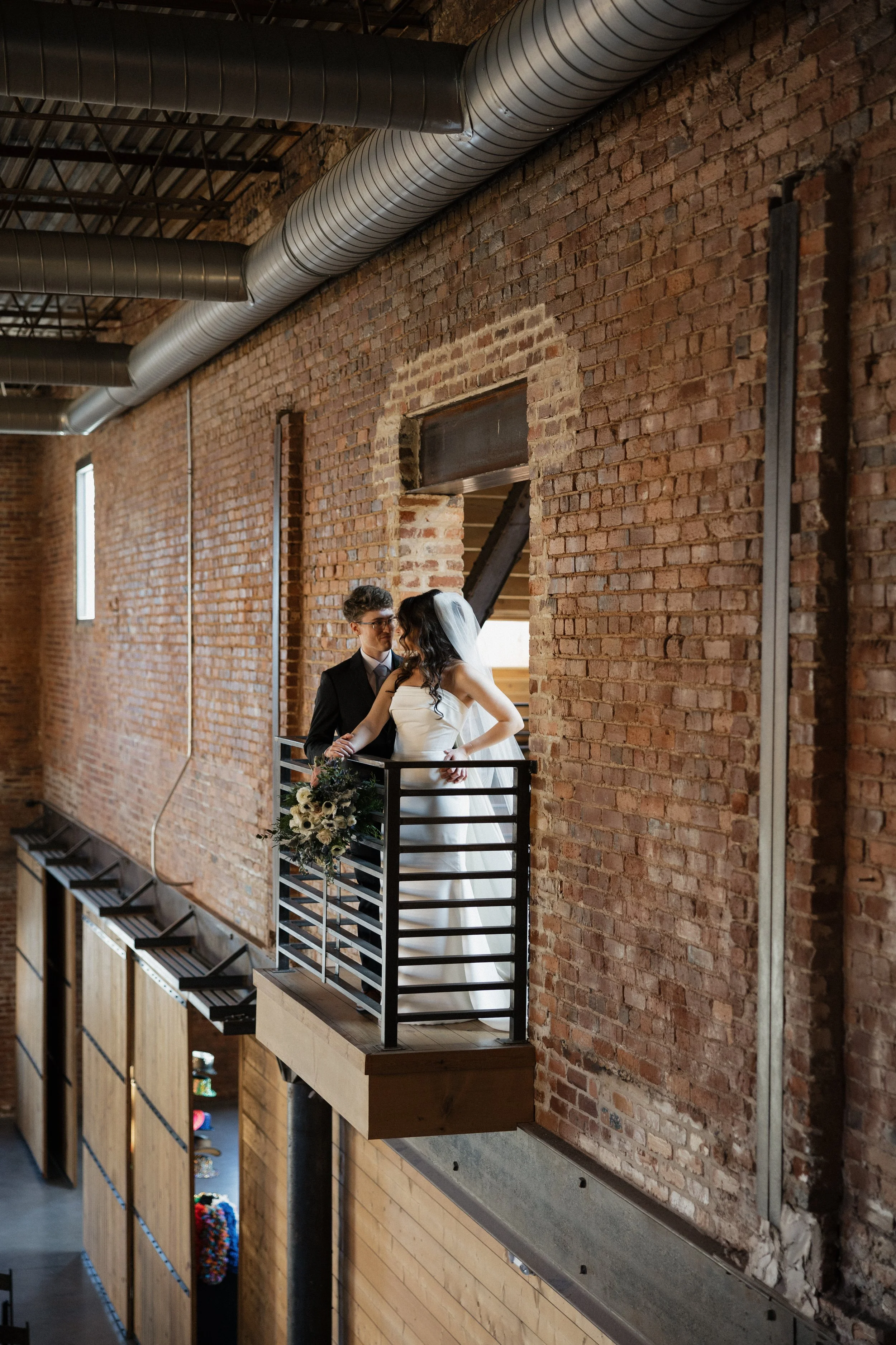Bride and groom standing on a small balcony with a brick wall background, during their wedding celebration.