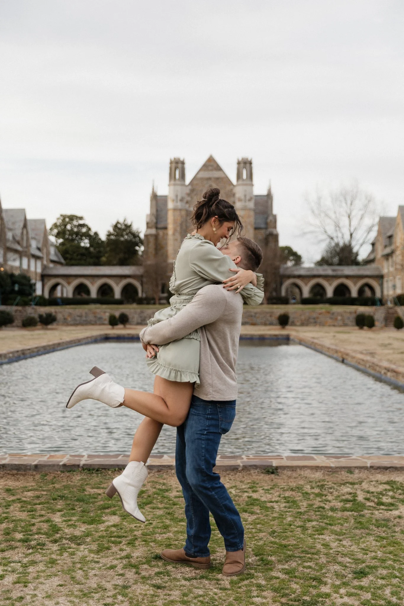 A couple is joyfully embracing outdoors near a pond, with a historic building in the background, as the woman is lifted into the air by the man.