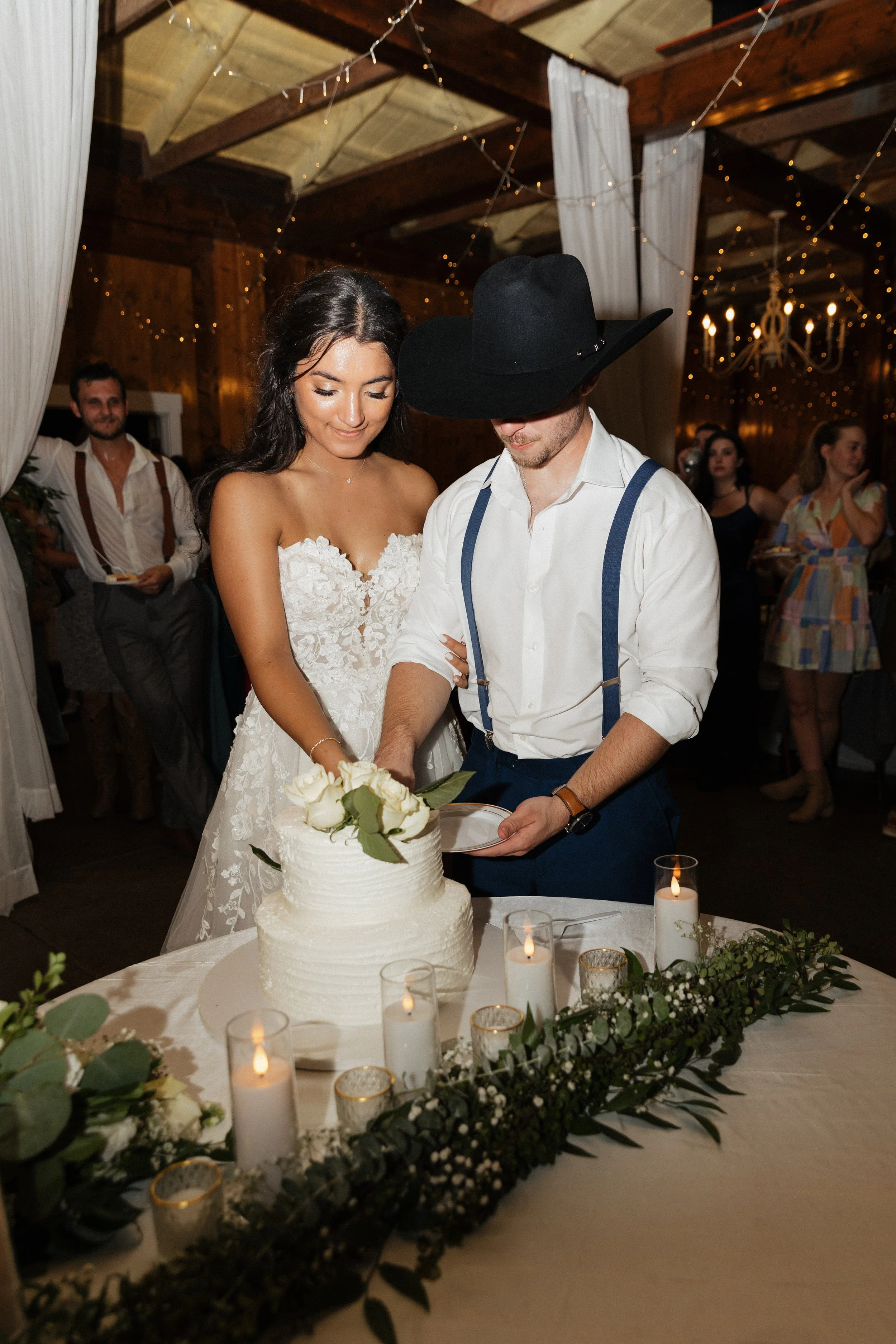 Bride and groom cutting a wedding cake together at reception with guests in background and candles on table.