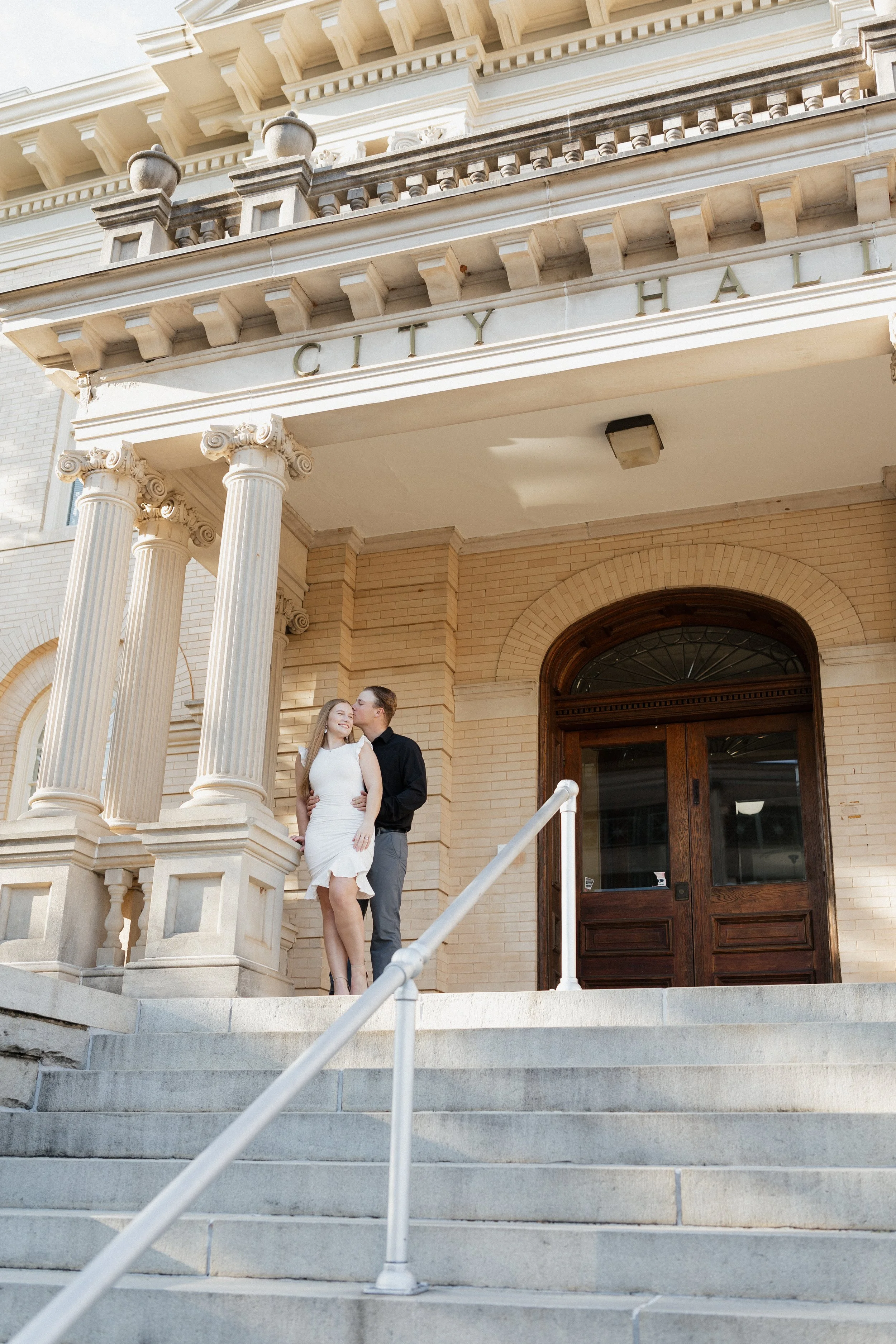 A couple standing on the steps outside a historic courthouse building, embracing and smiling, with the woman wearing a white dress and the man in a black shirt and gray pants.