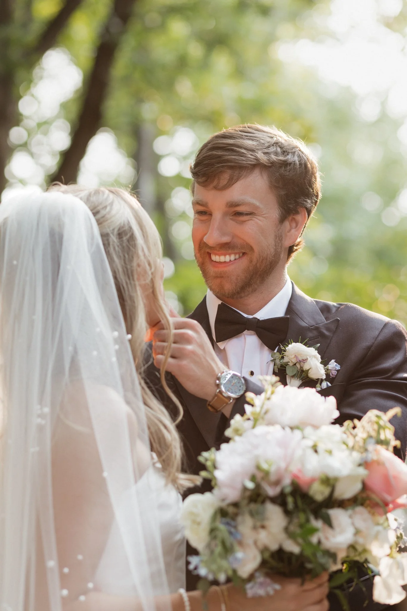 A groom smiling at a bride during a wedding ceremony outdoors, with green trees in the background, holding a bouquet of flowers.