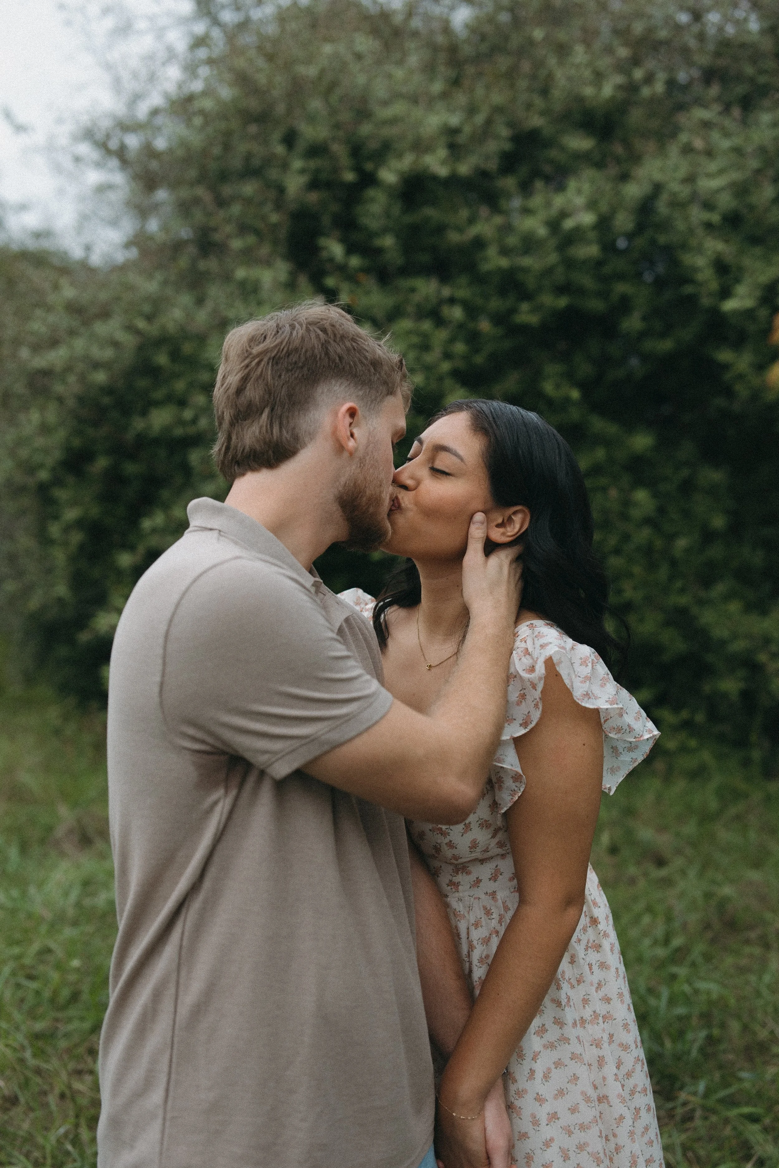 A couple kissing outdoors with trees and greenery in the background.