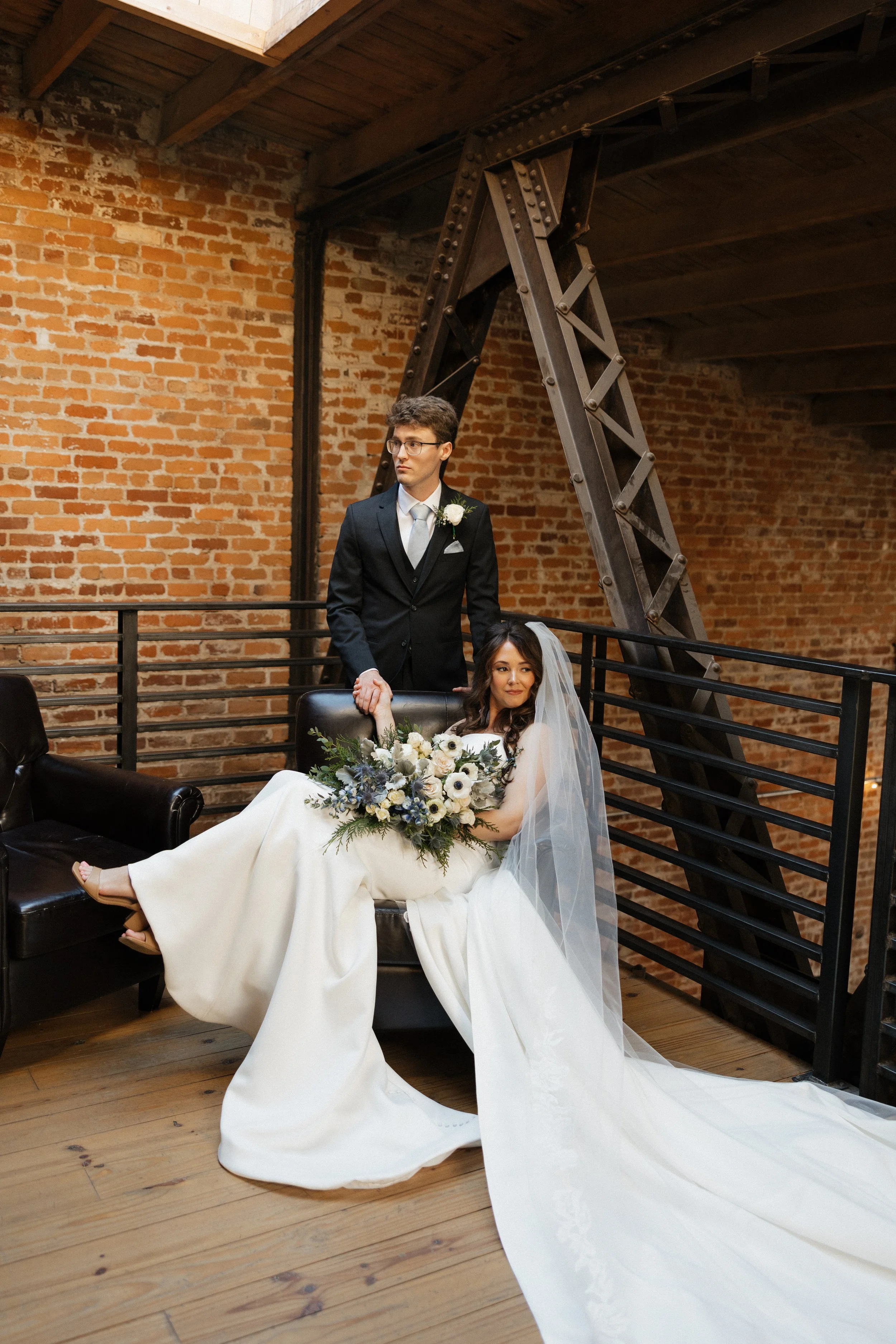 Bride sitting on a black leather couch, holding a bouquet of white and blue flowers, wearing a white wedding gown and veil, with a groom in a black suit and tie standing behind her, in an industrial-style venue with brick walls and metal staircase.