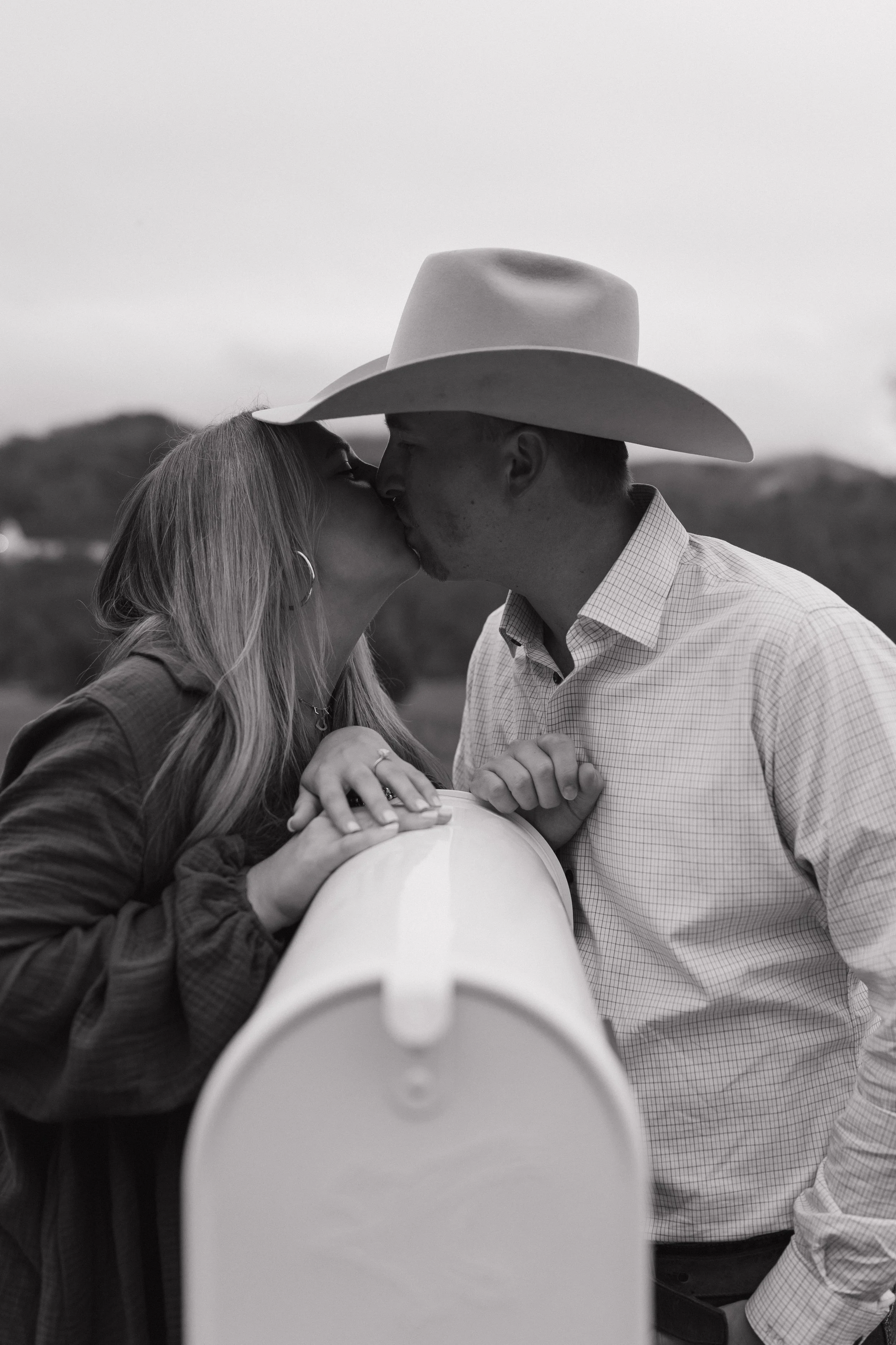 A black and white photo of a man and woman kissing, with the woman holding onto a mailbox and the man wearing a cowboy hat.