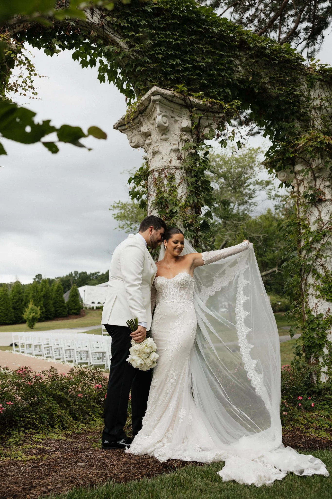 Bride and groom standing together outdoors under a stone archway with ivy, with the bride holding her veil and the groom holding a bouquet of white flowers.