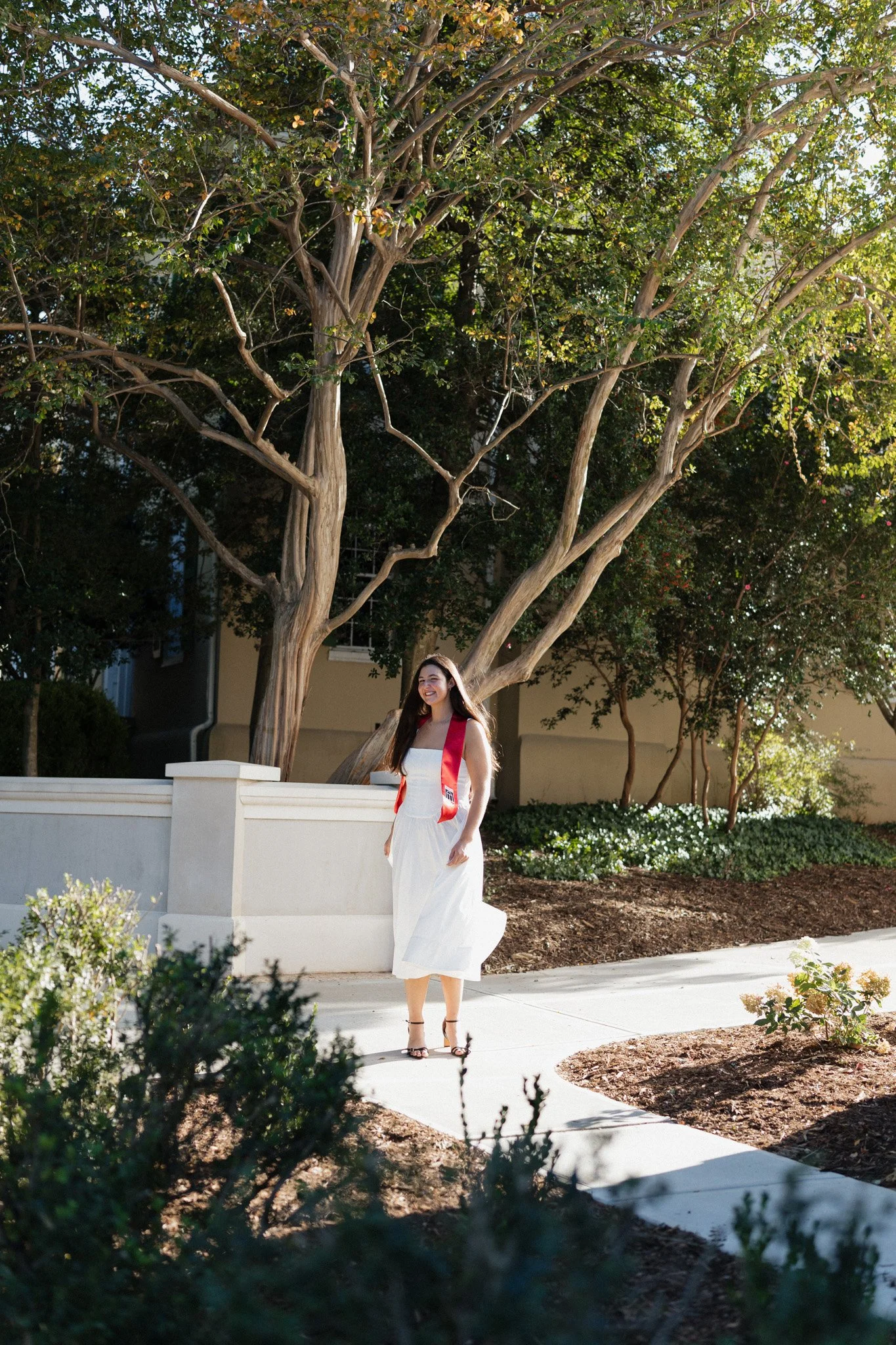 A woman in a white dress and black heels stands on a sidewalk. She has a red sash over her shoulders and is smiling. Behind her is a large tree with green leaves and a beige building.