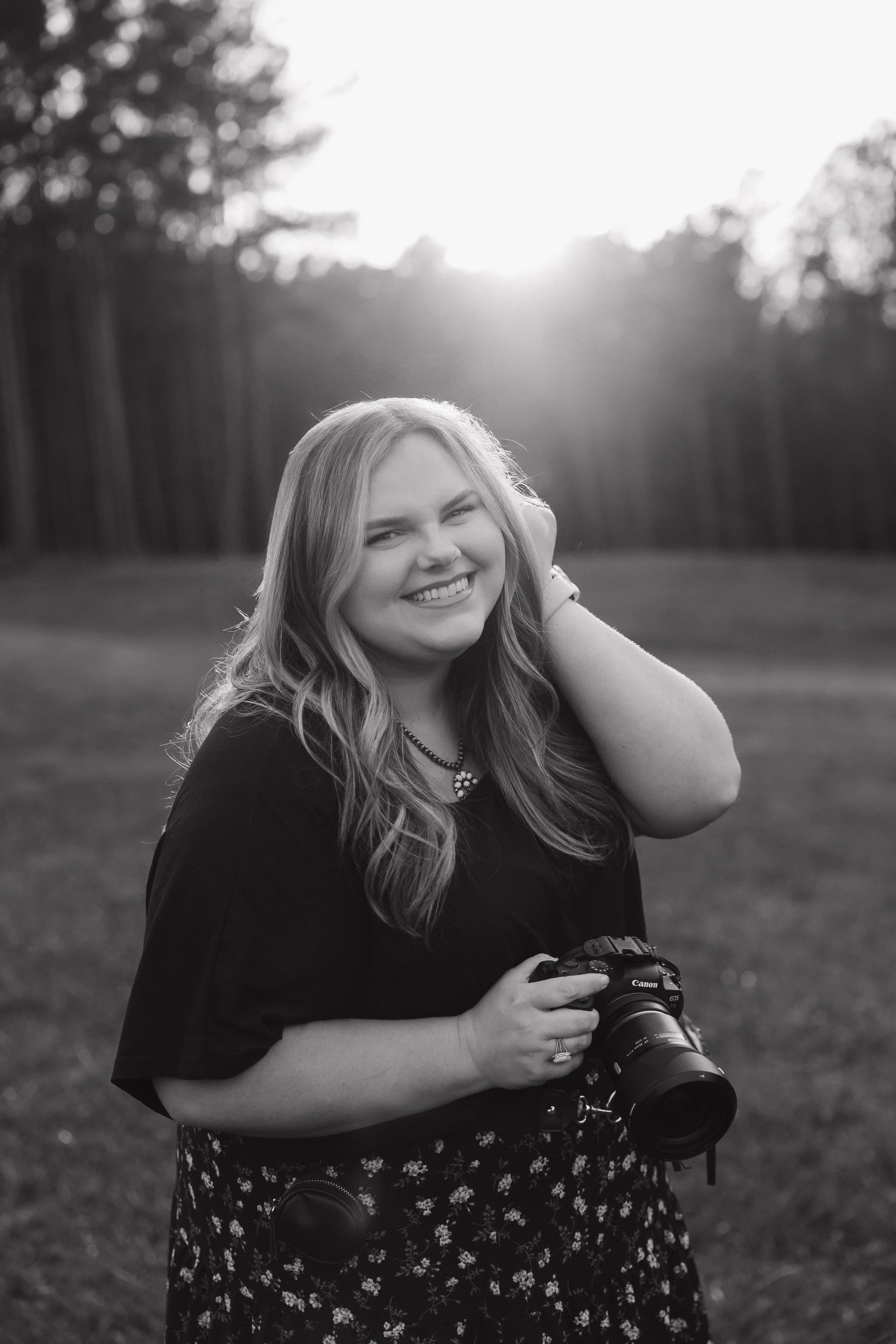 Black and white photo of a smiling woman holding a camera, standing outdoors at sunset with trees in the background.