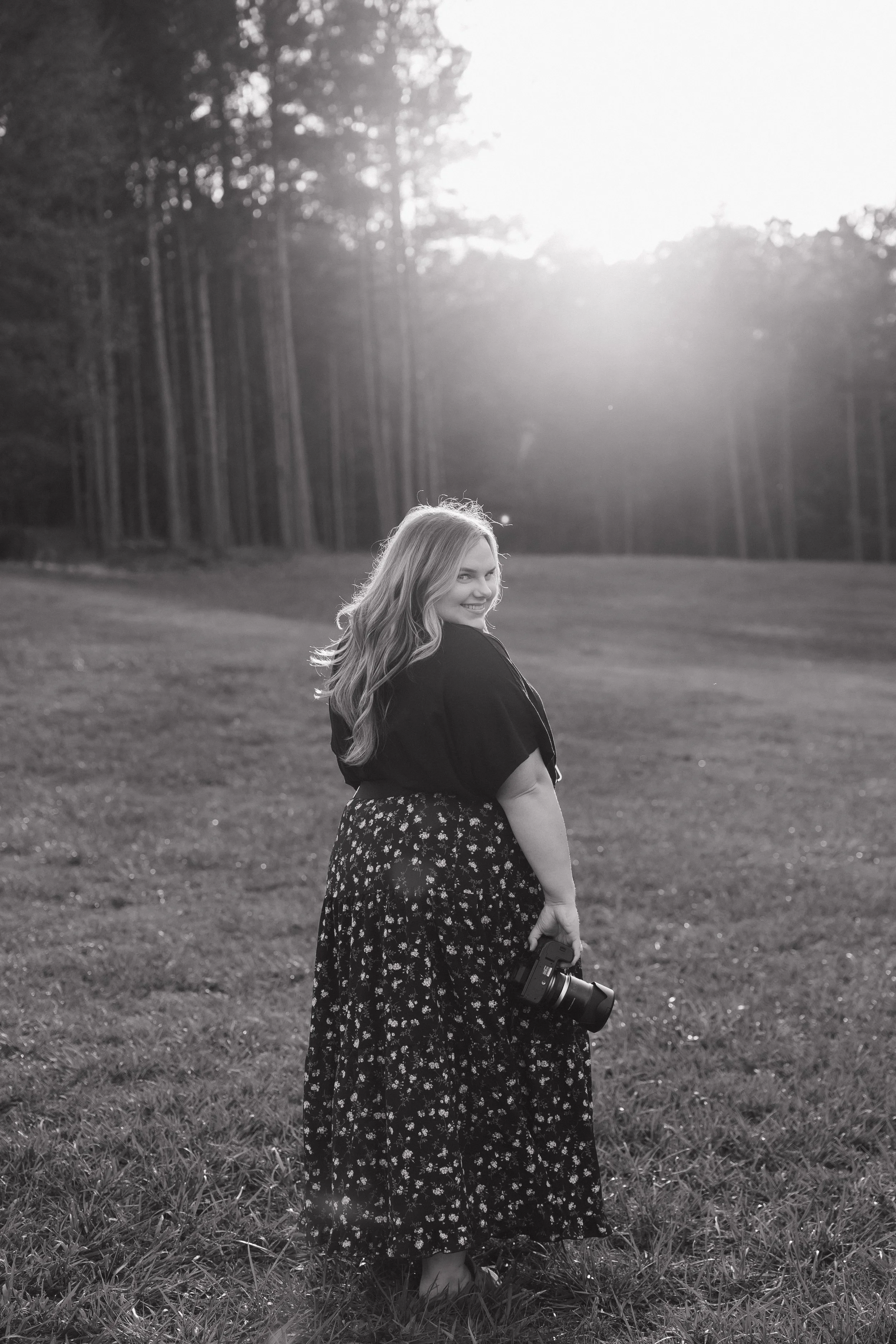 A woman with long, wavy hair stands on a grassy field holding a camera in her right hand, smiling and looking over her shoulder, with a backdrop of trees and sunlight.