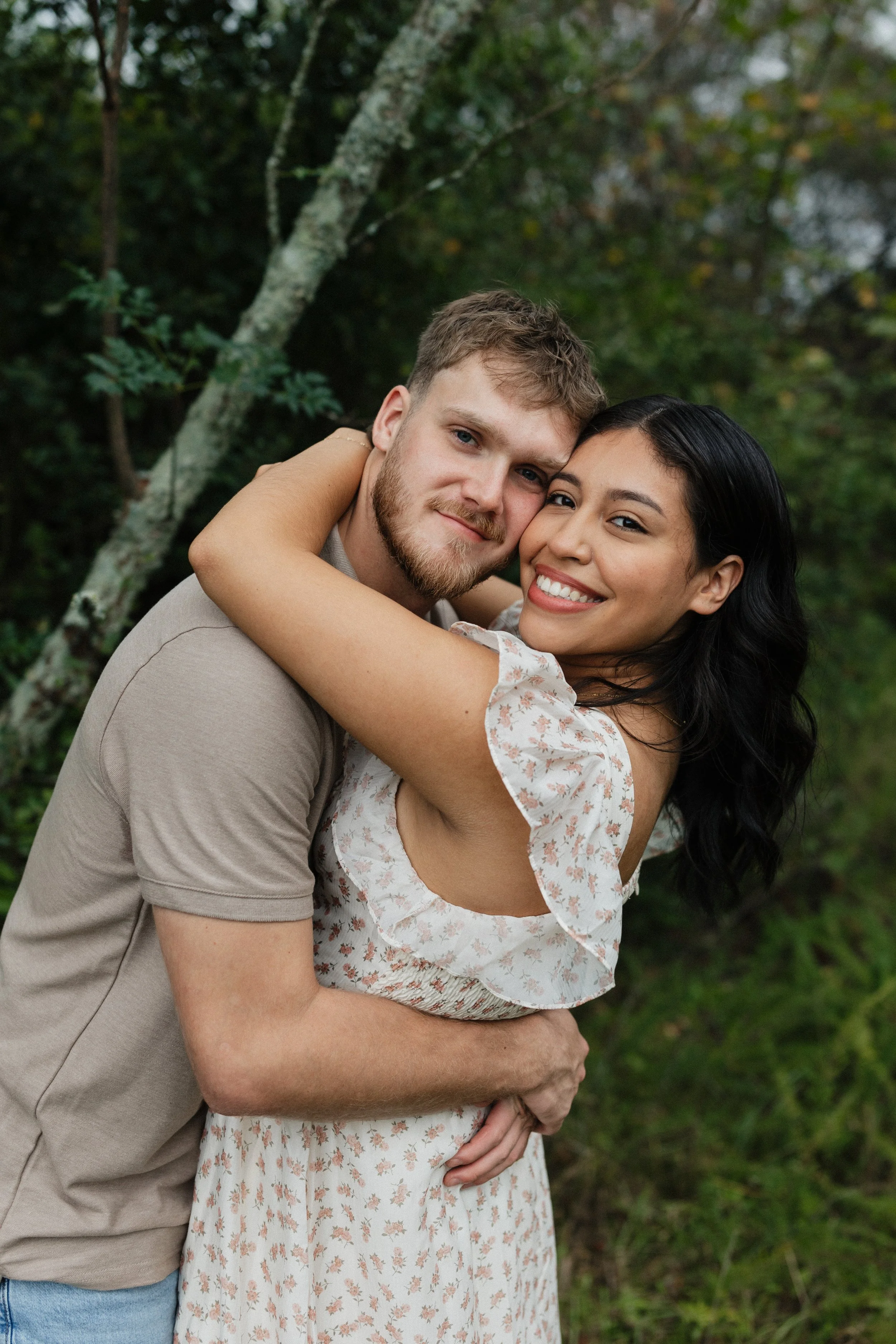 A happy couple embracing outdoors in a wooded area, smiling at the camera.
