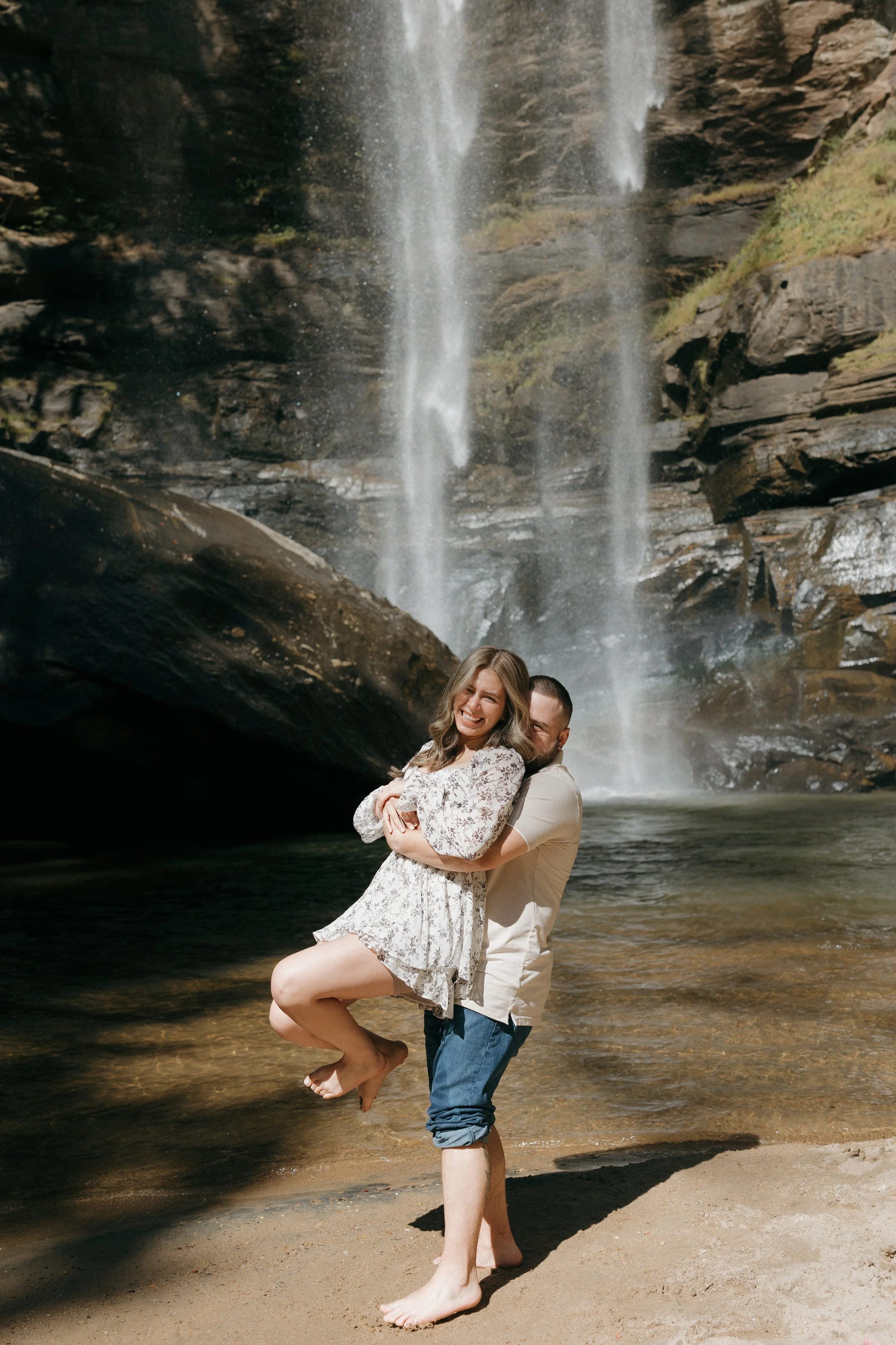 A happy couple standing on a sandy beach in front of a waterfall, with the man lifting the woman in his arms. The woman is smiling and wearing a floral dress, while the man is dressed casually in a light shirt and rolled-up jeans.