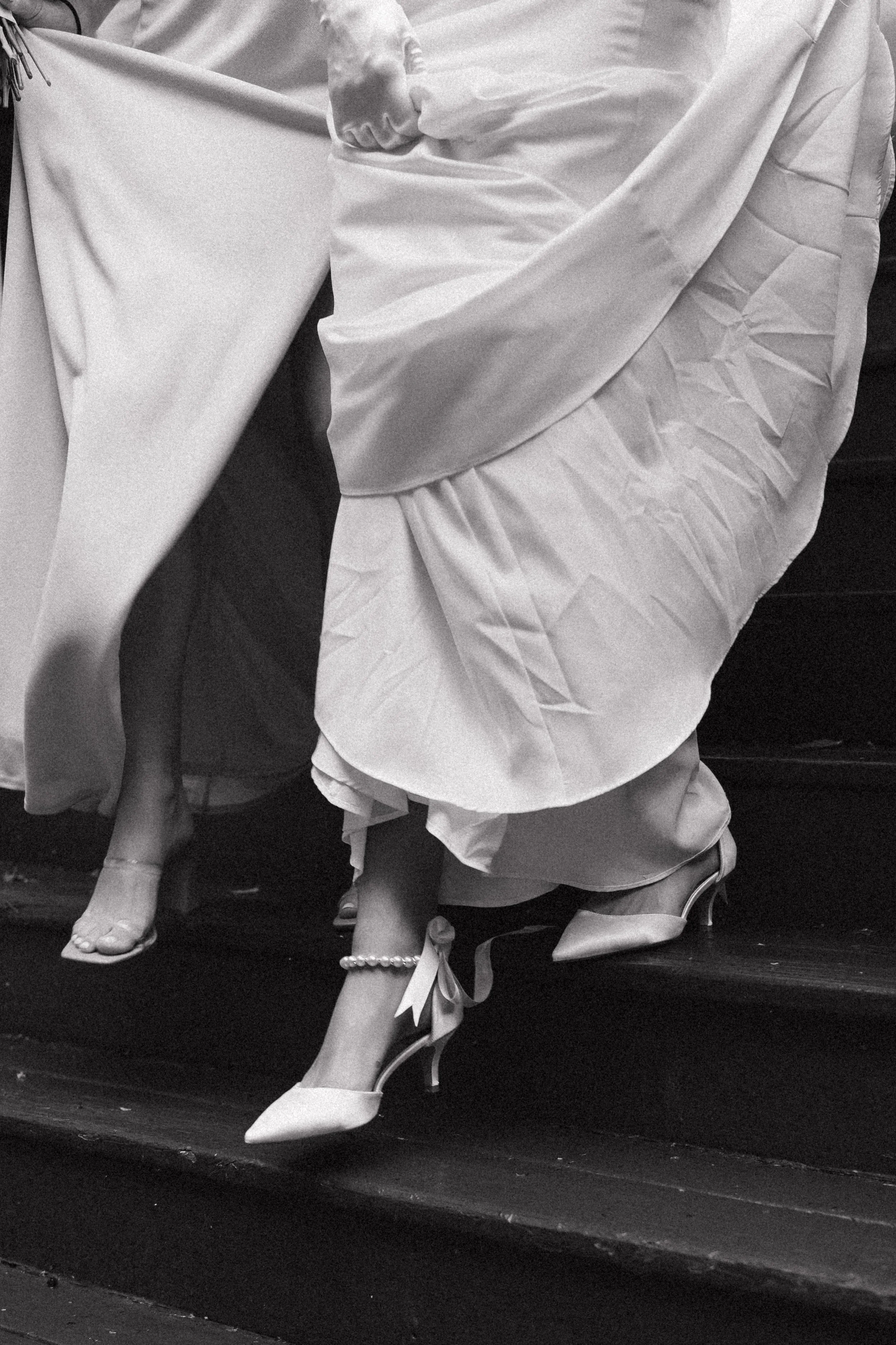 Close-up of women's feet in high-heeled shoes, one with a pearl anklet and bow, on a staircase. The women are wearing formal dresses.