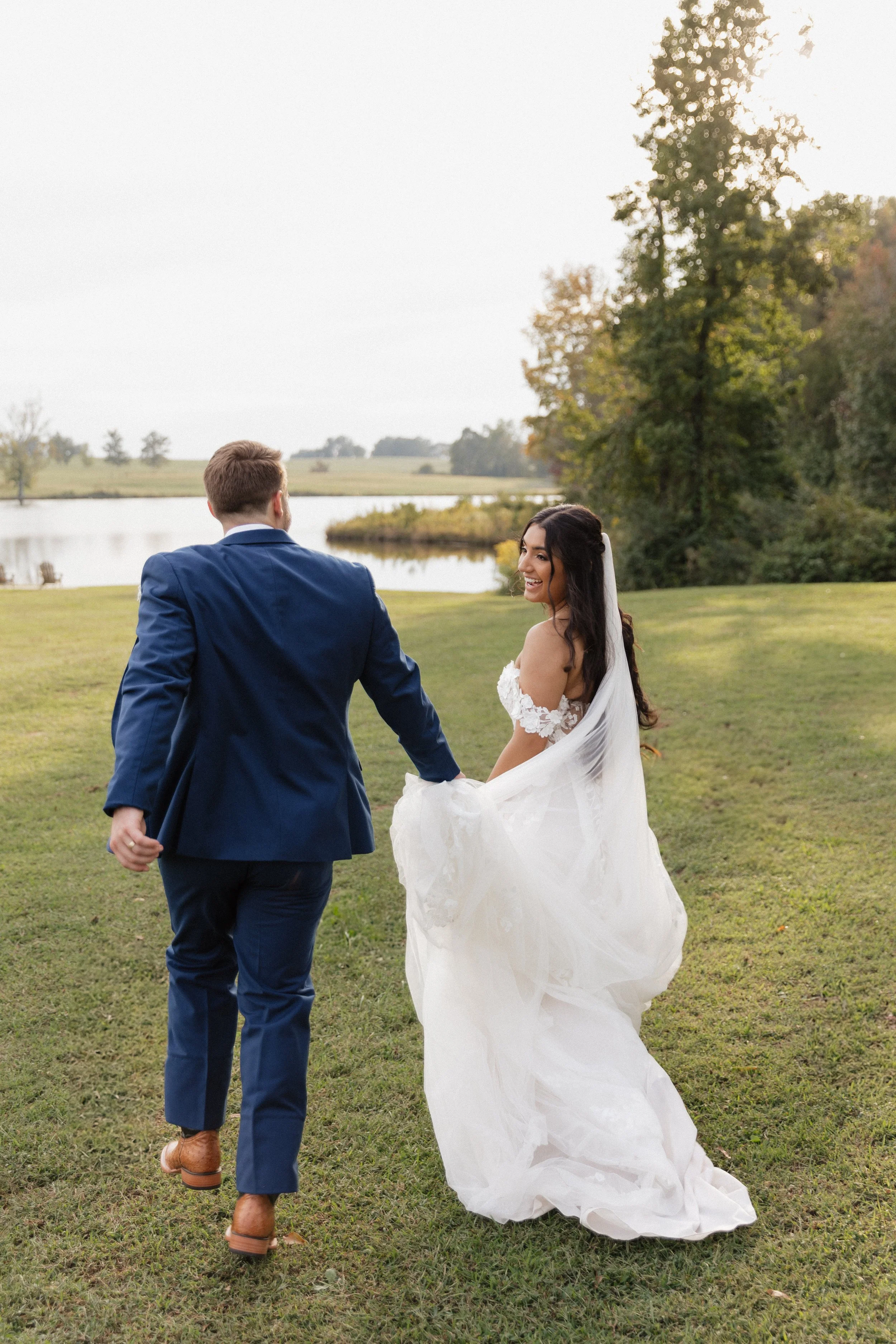 A newlywed couple walking hand-in-hand outside near a lake with trees in the background, the bride in a white wedding dress and veil, and the groom in a blue suit.