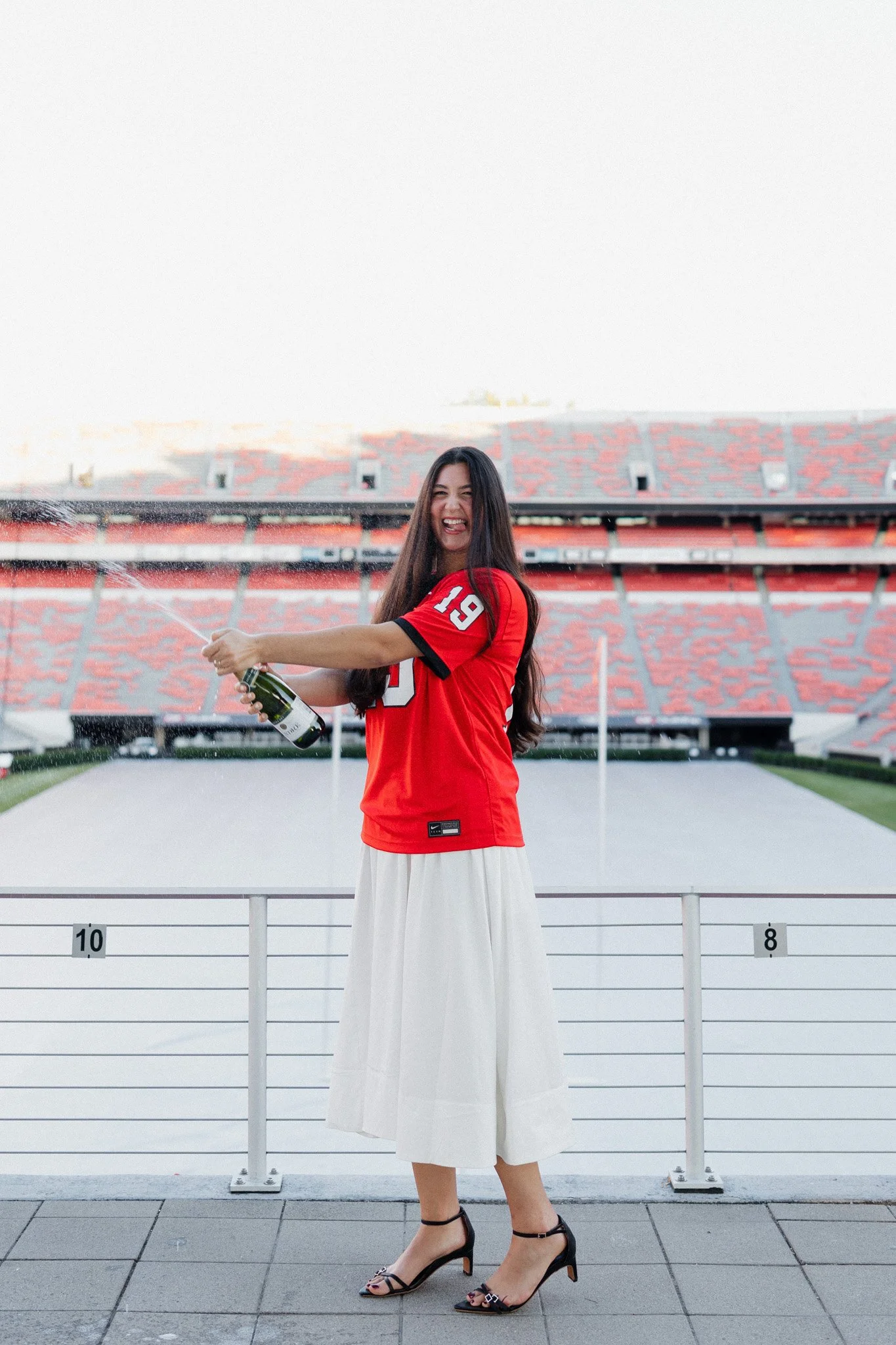 A woman in a red sports jersey and white skirt celebrating on a stadium balcony while spraying champagne.