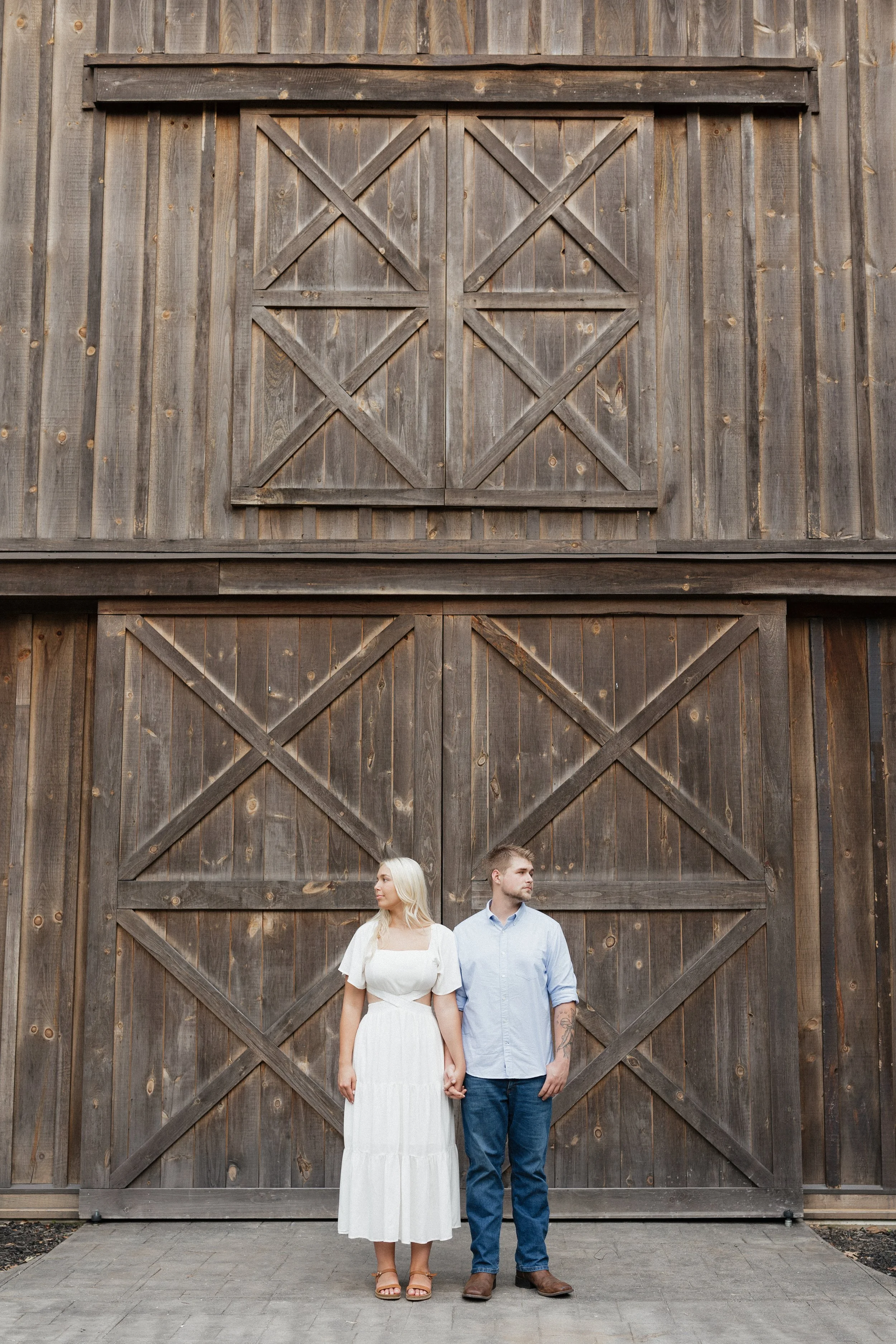 A couple standing hand-in-hand in front of a large wooden barn door, holding hands and looking in opposite directions.