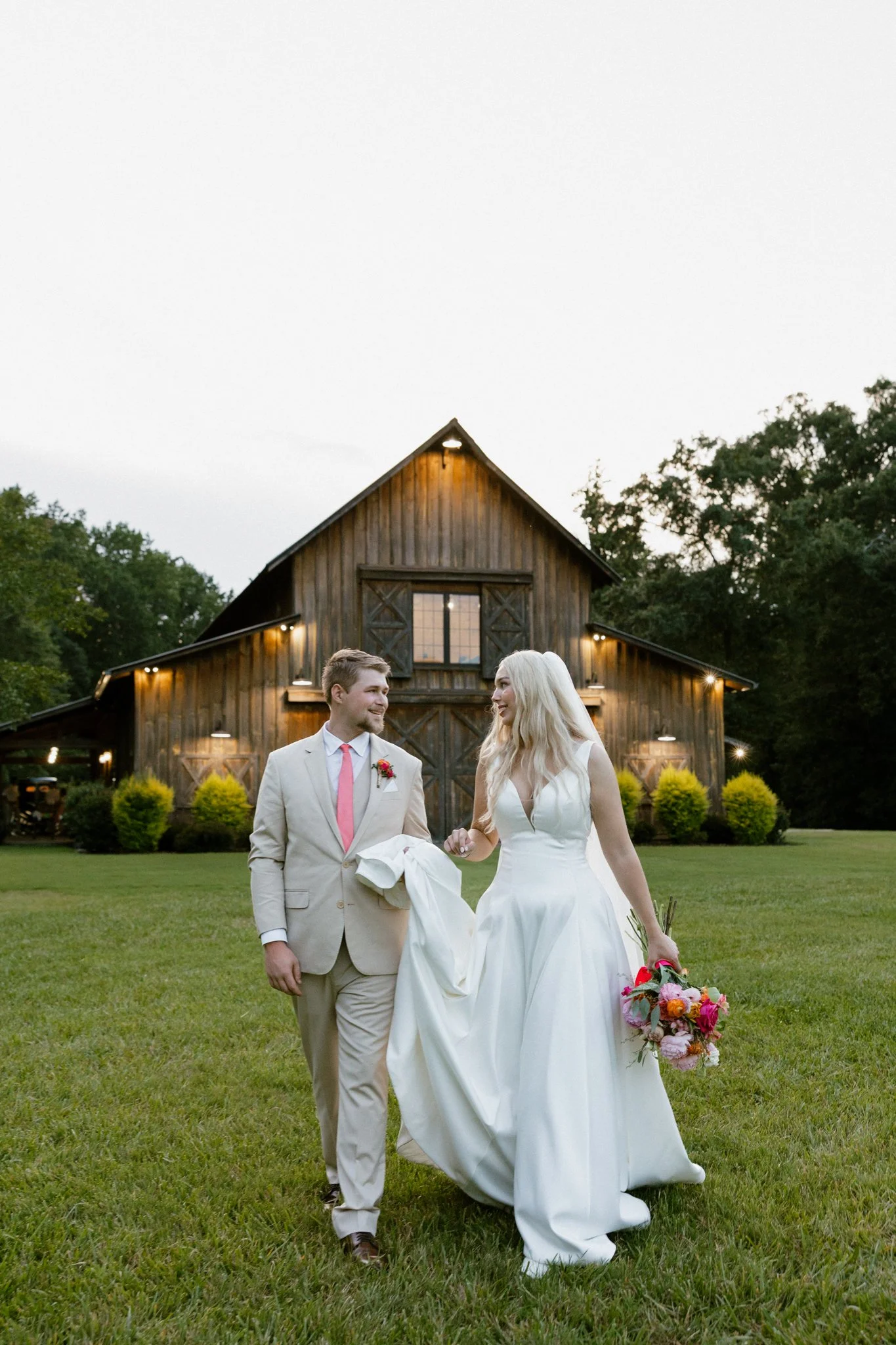 A bride and groom walking on grass in front of a rustic wooden barn at dusk, smiling at each other with the bride holding a colorful bouquet.