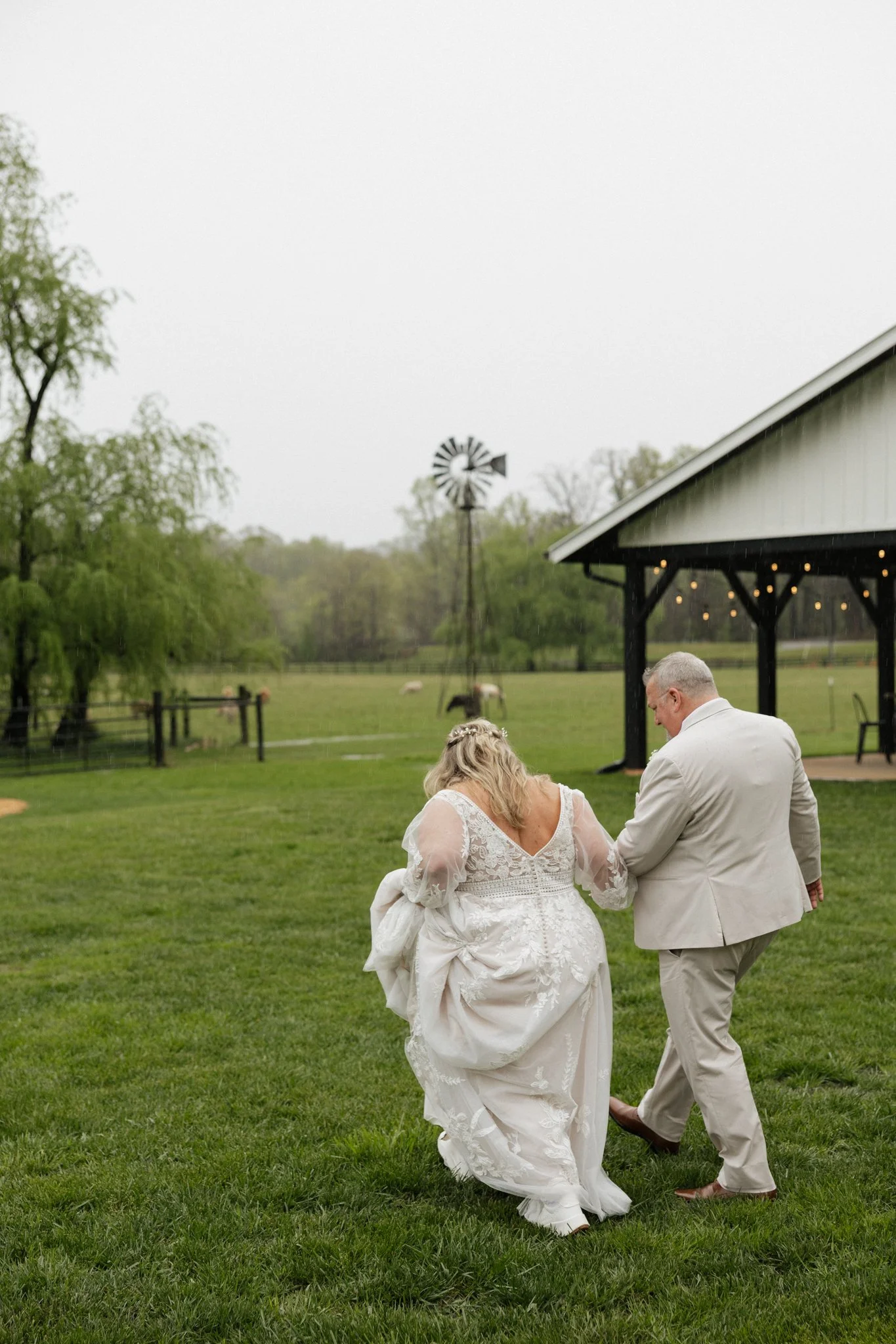 A bride and groom holding hands walking on a grassy field near a barn on a farm during a rainy day.