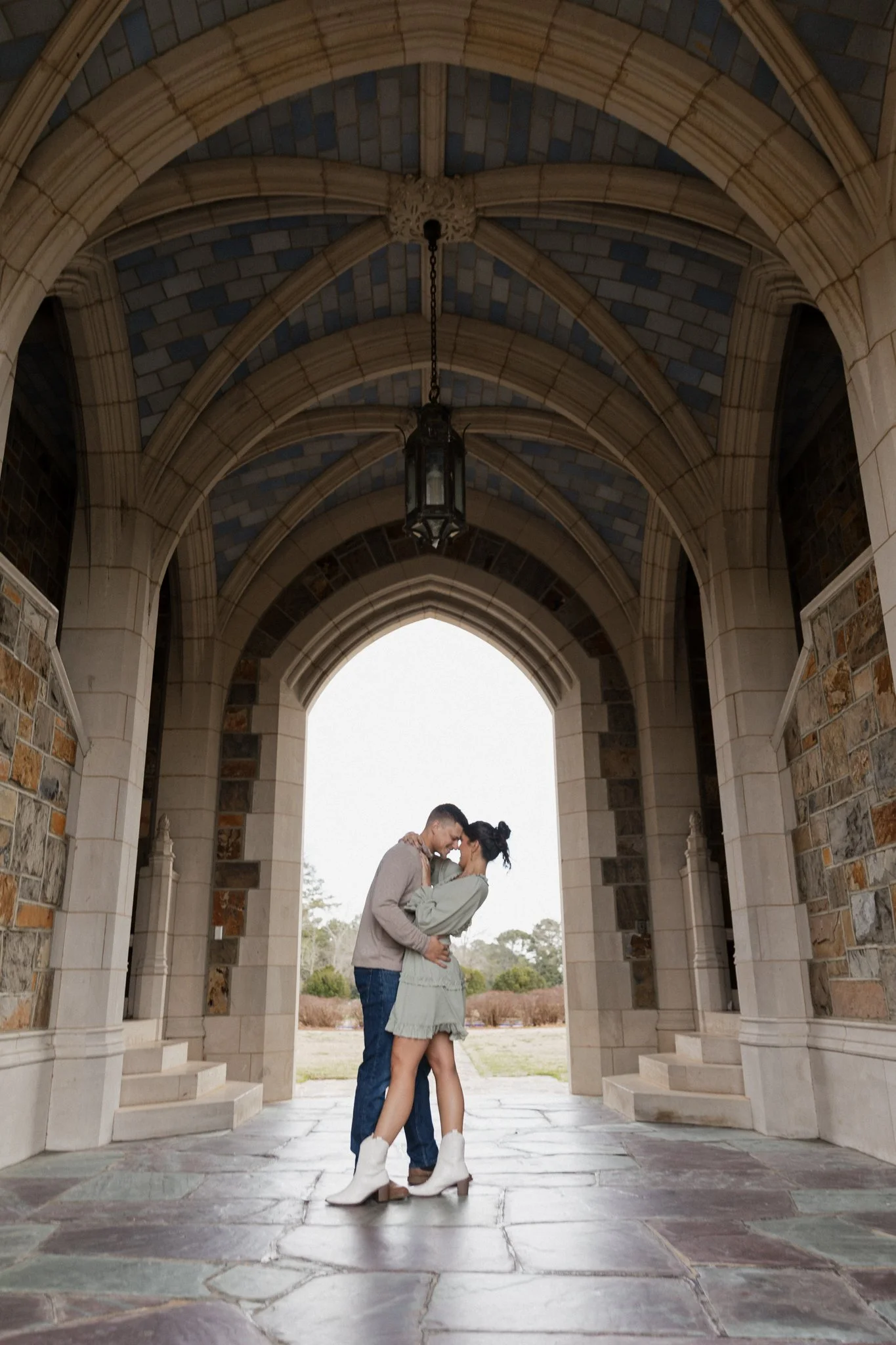 A couple embraces, standing under a stone archway with ornate ceiling and hanging lantern, outdoors in front of a bright open landscape.
